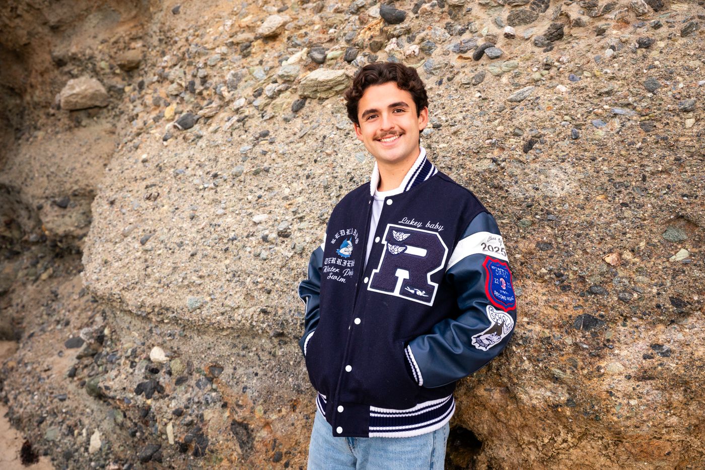 Graduate poses near rocky coastal formation wearing varsity jacket. Beach photography, Huntington Beach, Laguna Beach, Redlands photographer