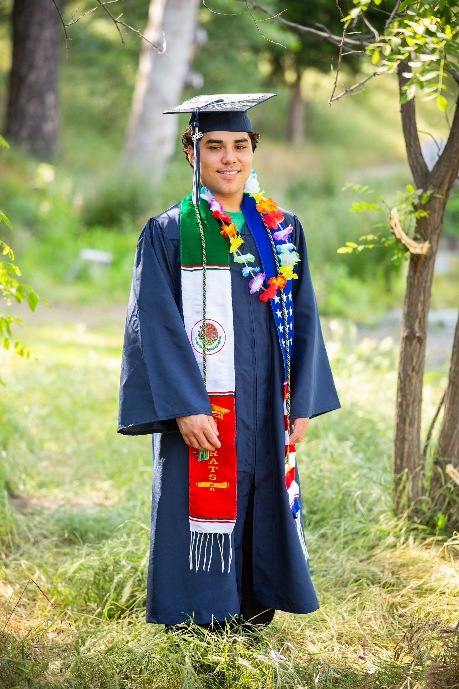 Graduate dressed in navy blue cap and gown with colorful leis and graduation stoles poses outdoors in nature setting.