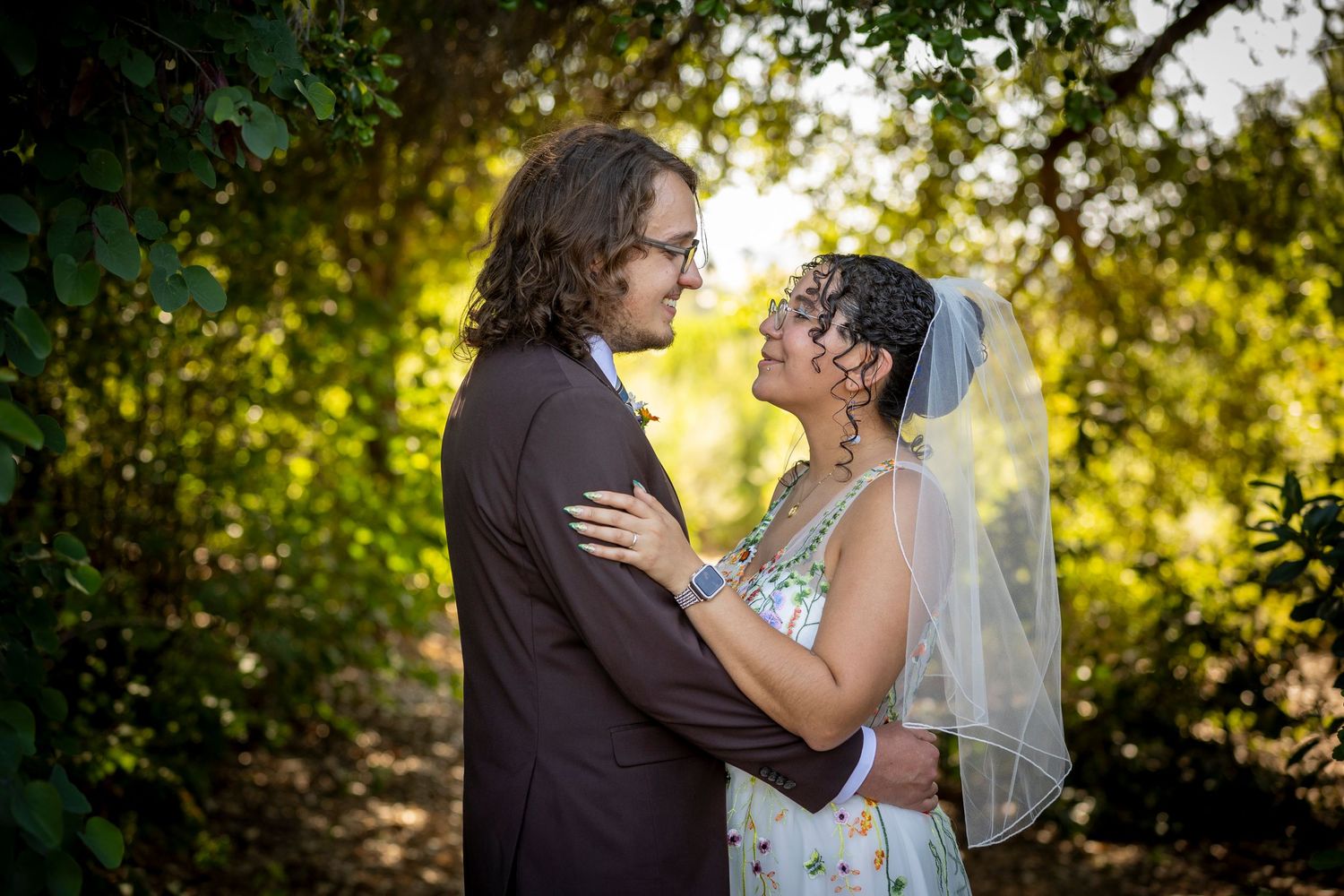 Two people embrace in a romantic outdoor setting with sunlit trees providing a natural backdrop.