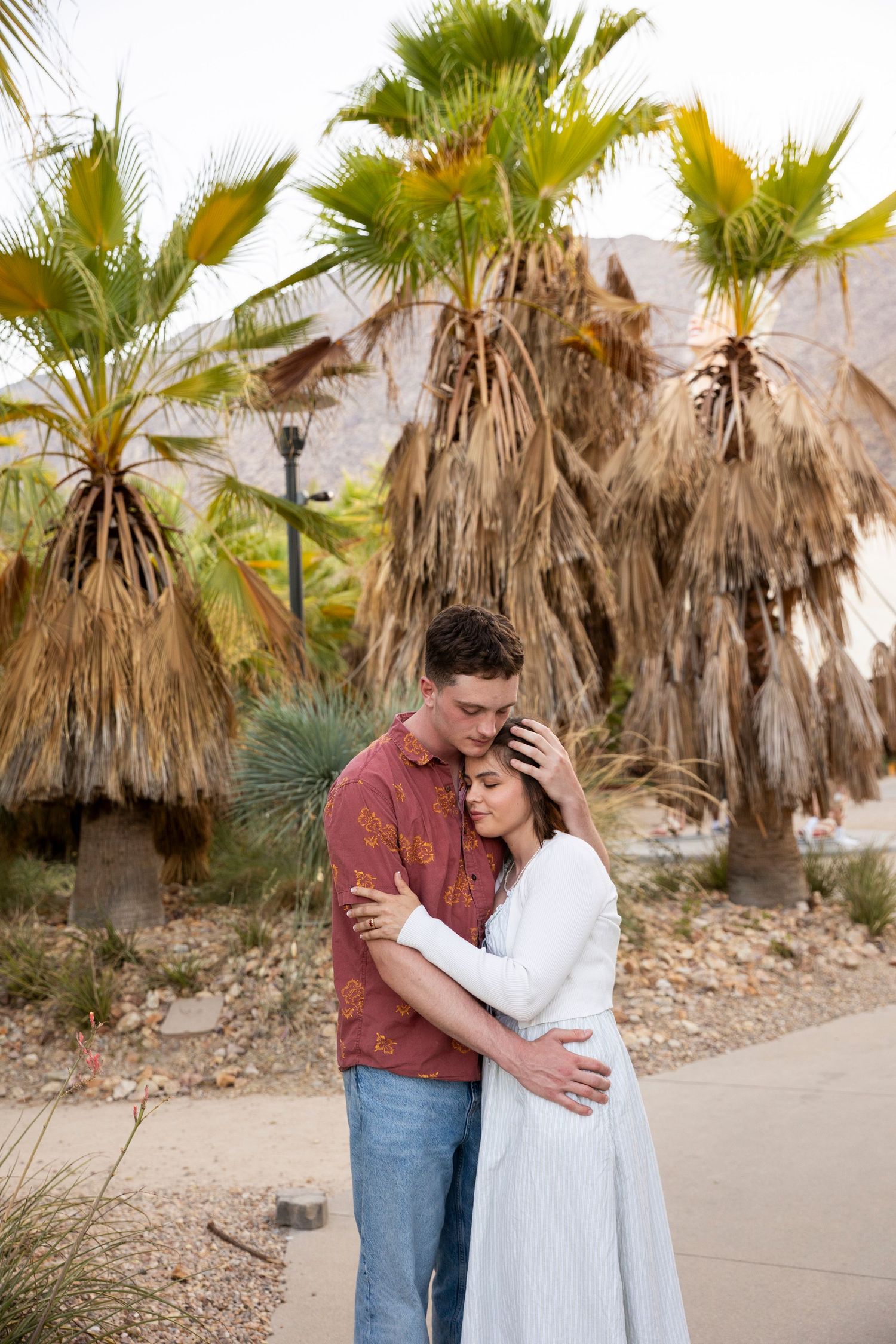 A couple shares a tender embrace while standing on a path surrounded by desert palm trees.