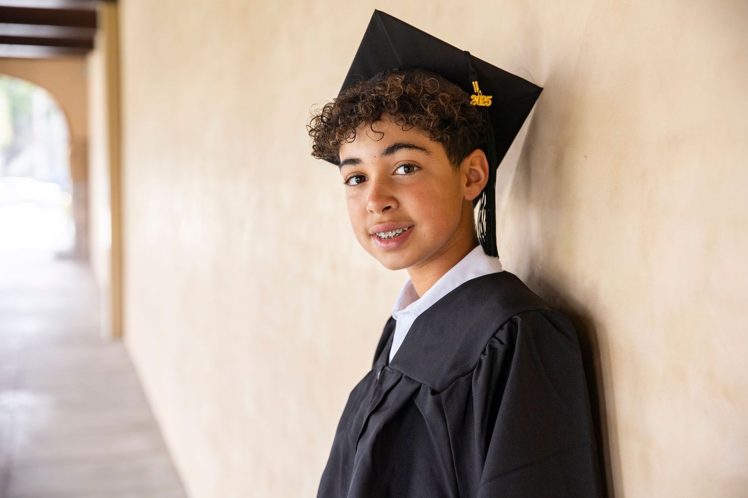 A graduate in black cap and gown smiles while standing in a hallway.