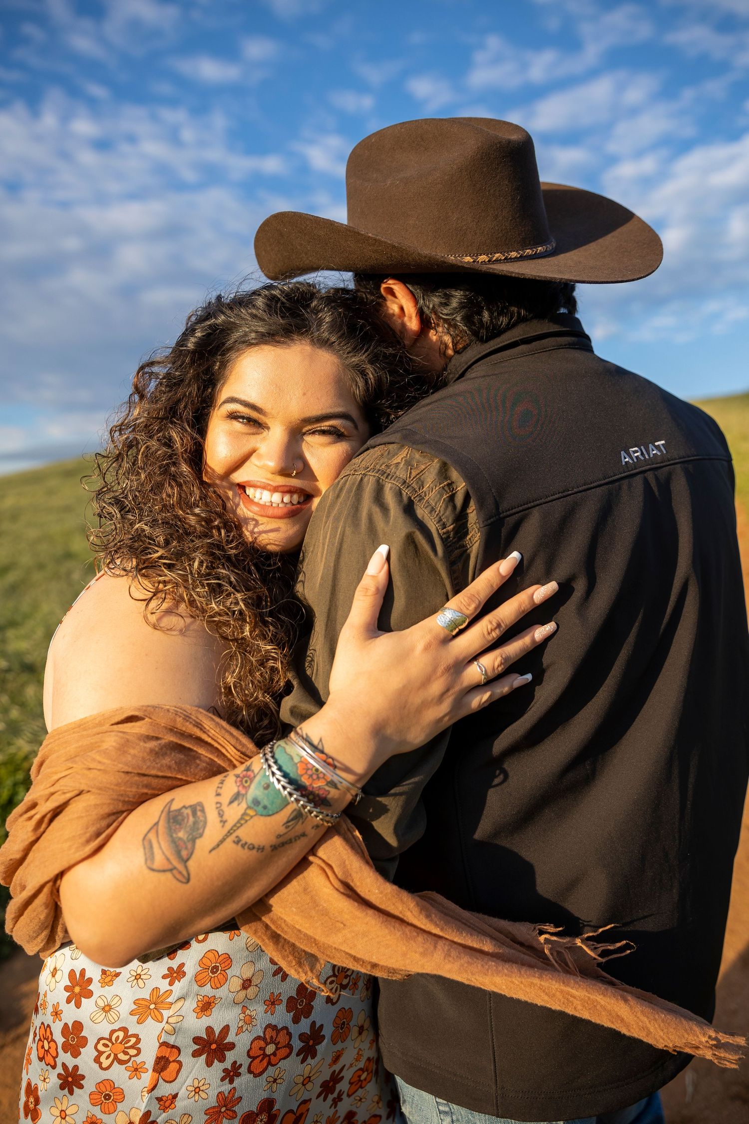 A couple embraces outdoors with one wearing a cowboy hat and brown jacket while the other smiles in a floral dress.