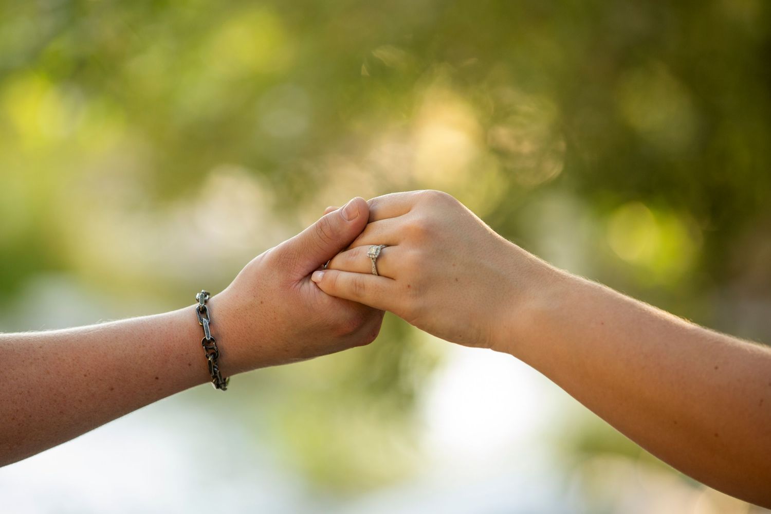 Two hands gently clasp together against a soft bokeh background of green and white in natural sunlight.