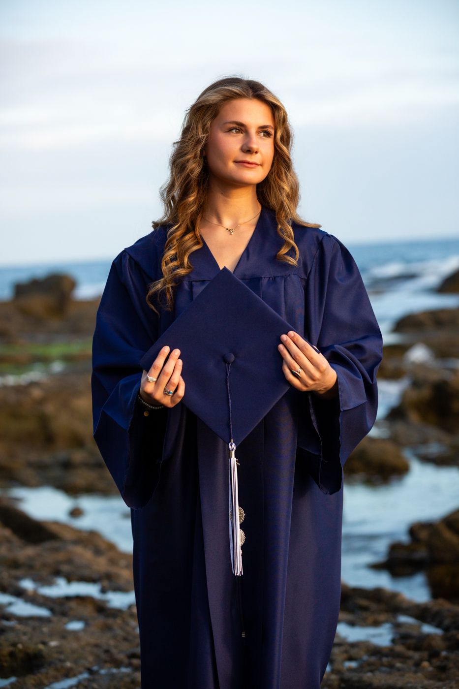 Graduate in dark blue gown poses with tassel against rocky coastline. Beach photography, Huntington Beach, Laguna Beach, Redlands photograph