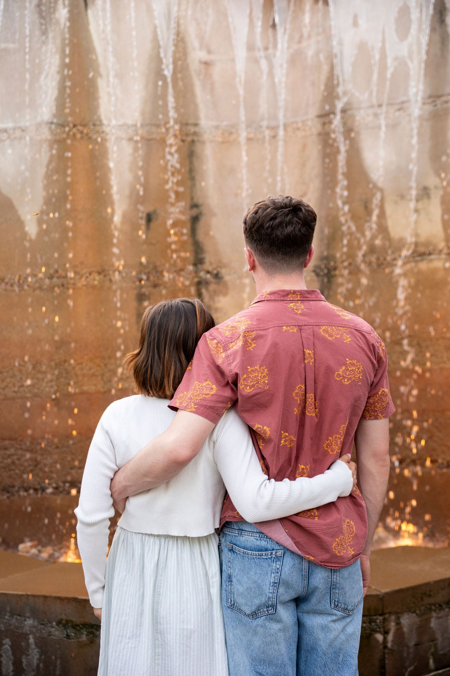 A couple in embrace standing against a rusty metal wall, with fairy lights twinkling in the background.