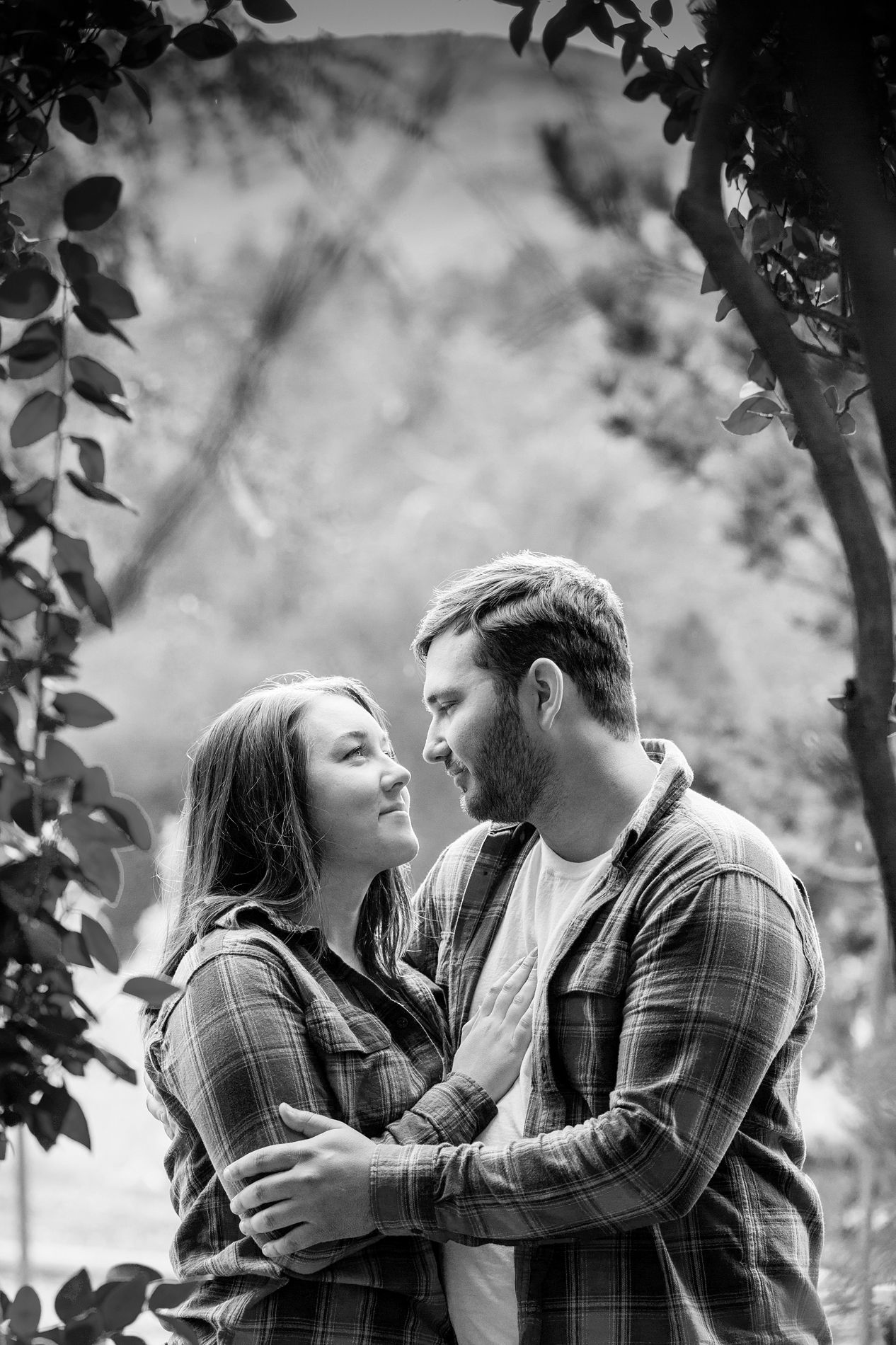 A romantic black and white portrait shows a couple in matching plaid shirts embracing outdoors among tree leaves.