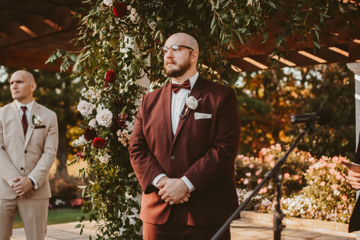 Groom in burgundy suit with bow tie stands at outdoor wedding altar adorned with red and white floral arrangements.