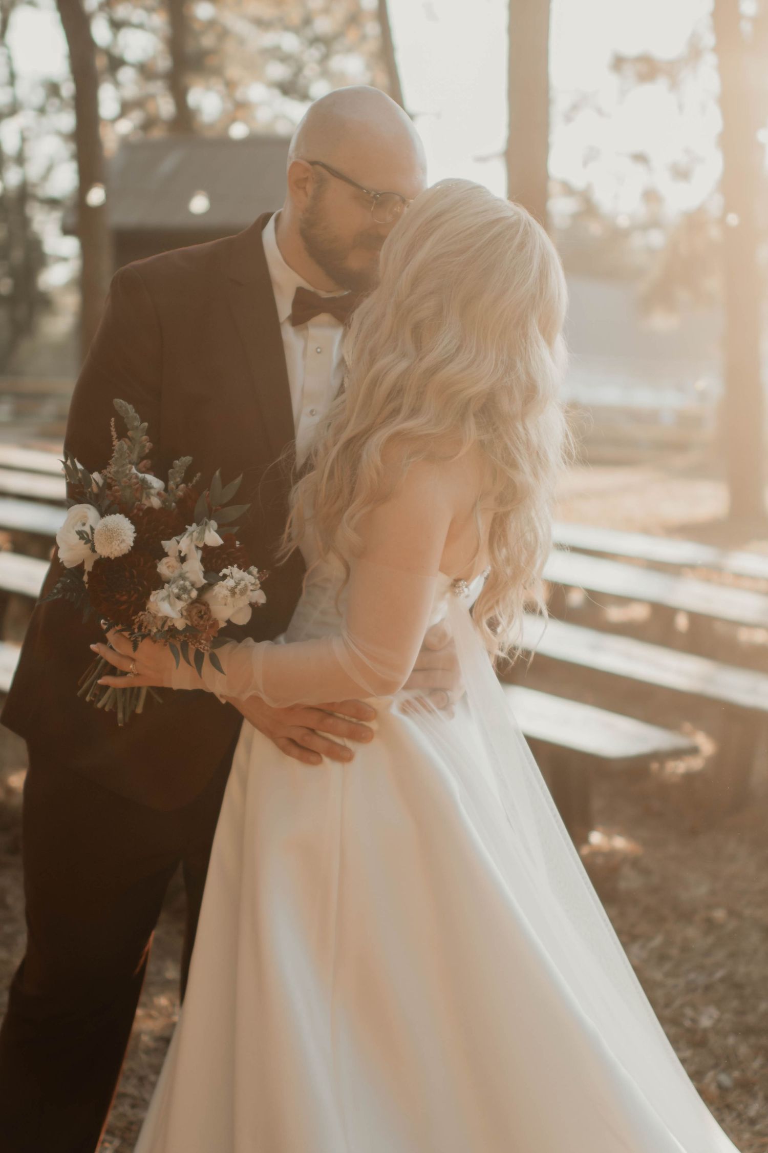 A bride and groom share a romantic embrace outdoors in golden sunlight, she holds a white and burgundy bouquet.