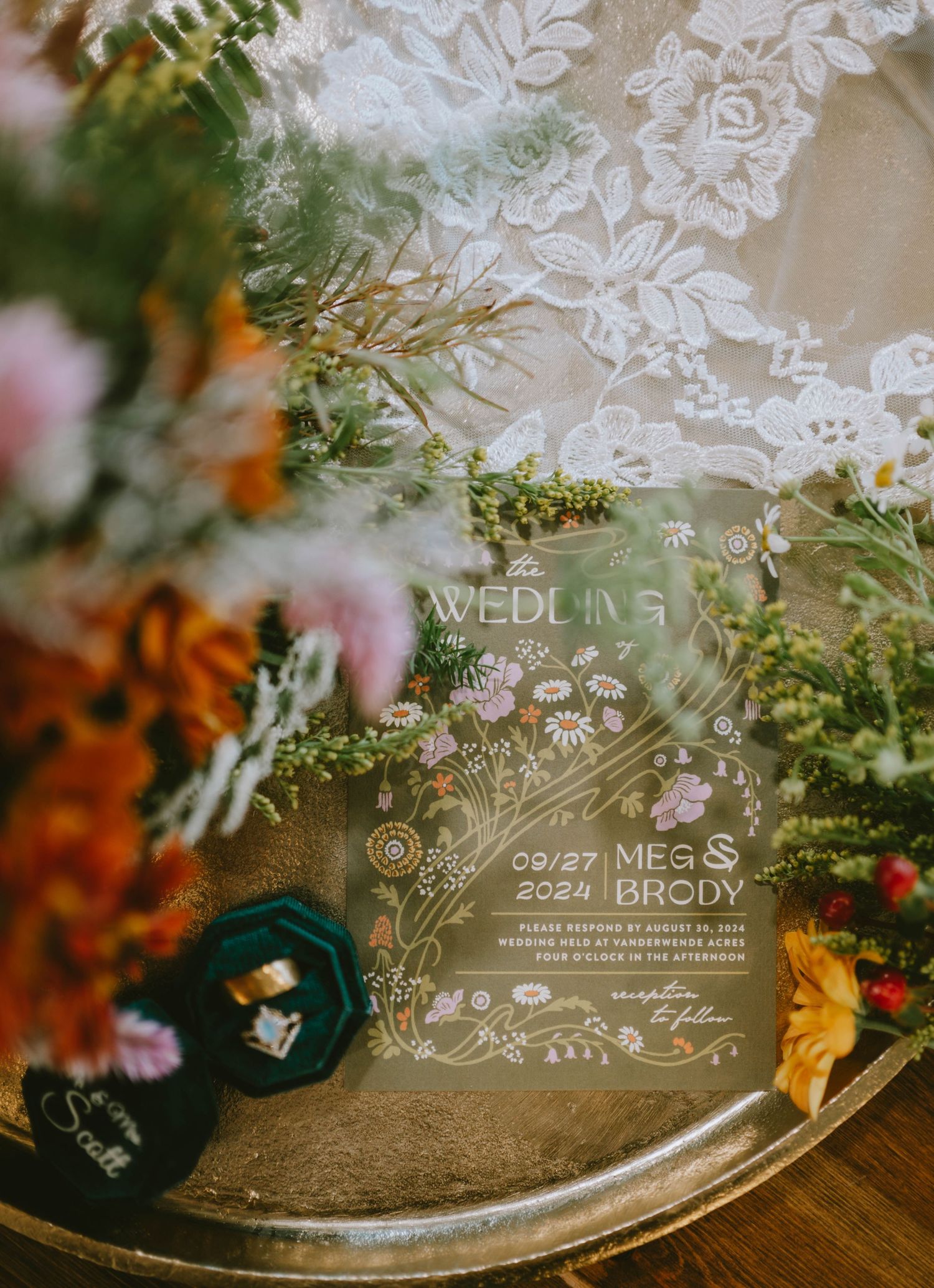Acrylic wedding invitation for Meg and Brody dated 09/27/2024, surrounded by wildflowers and lace on a silver tray.