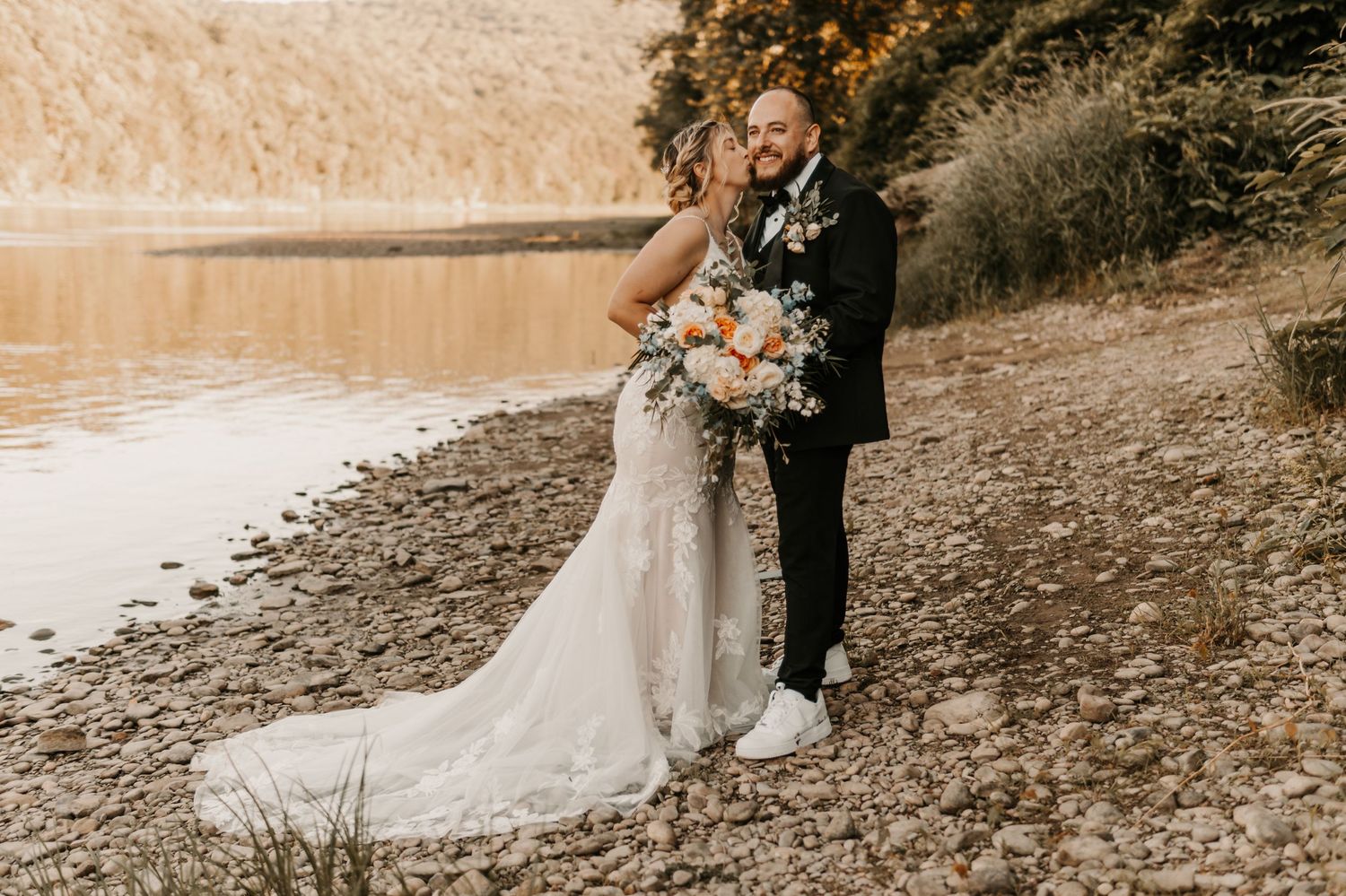 Bride and groom embracing on rocky lakeside shore, bride holding floral bouquet in white lace gown.
