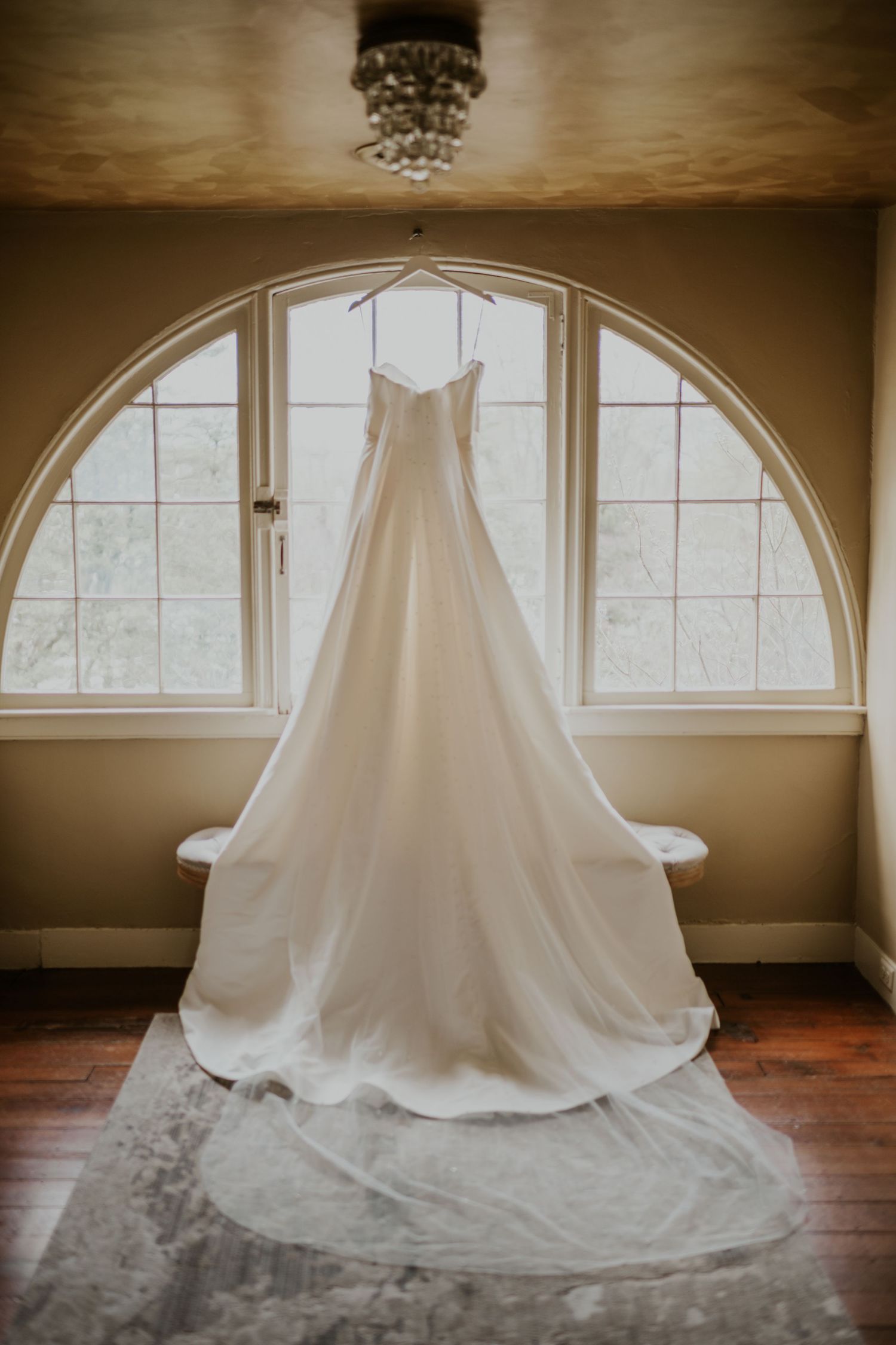 Elegant white wedding dress with a long train hanging on a hanger in front of an arched window in a warmly lit room.
