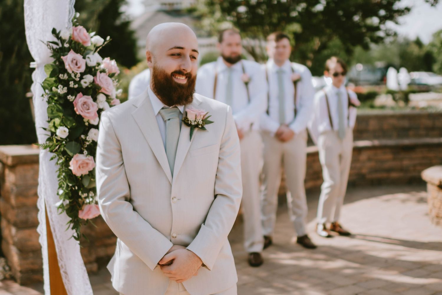 Smiling bald groom with beard in beige suit stands at wedding altar with groomsmen and floral arch.