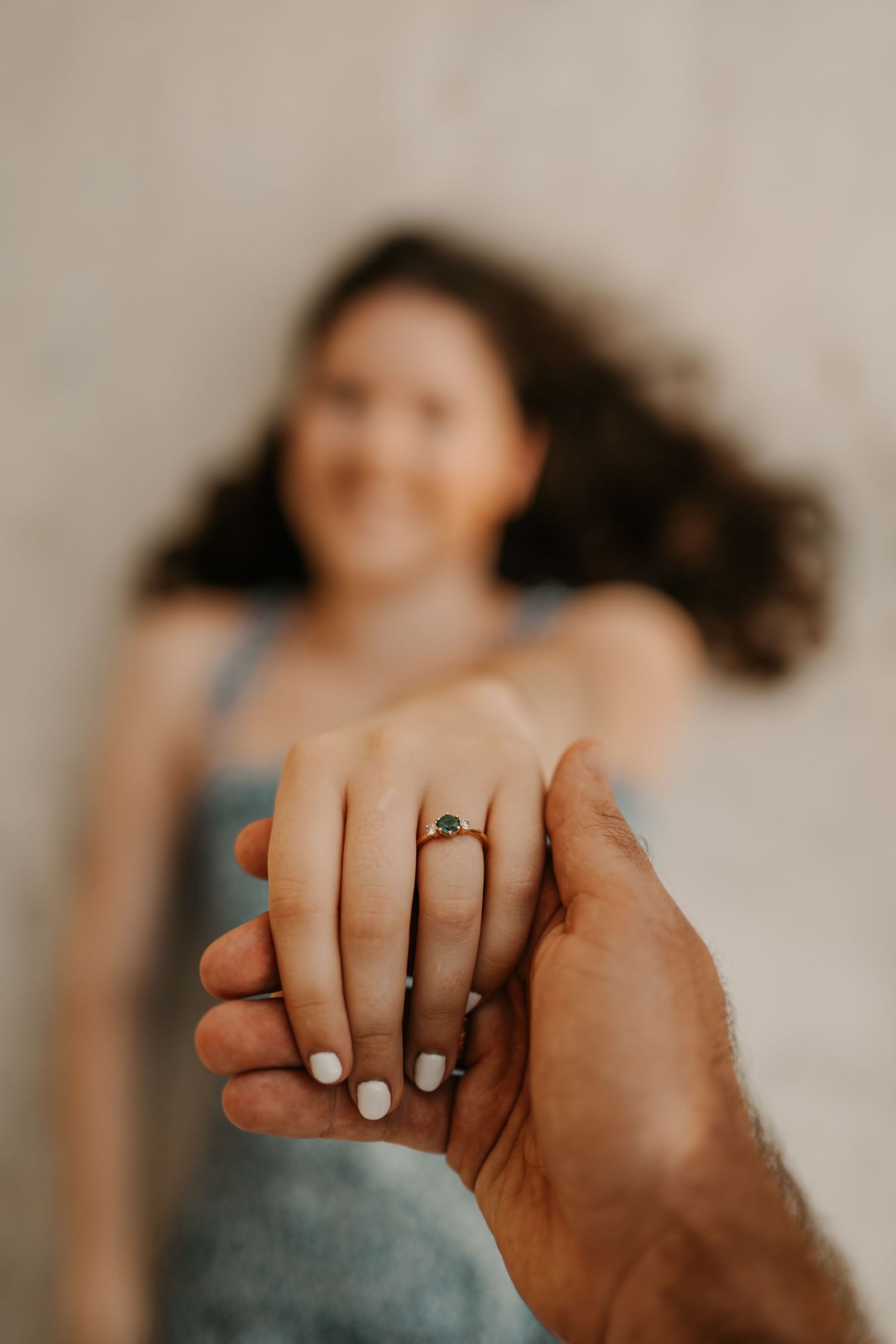A smiling woman holds out her hand displaying a gold engagement ring with a green emerald gemstone.