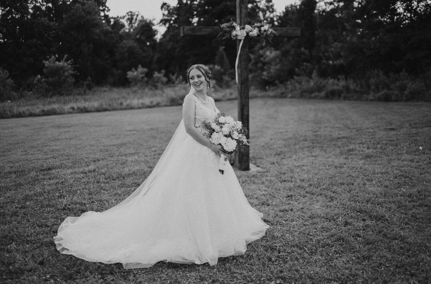Black and white photo of a bride in a flowing ball gown holding a bouquet, standing on a green lawn.