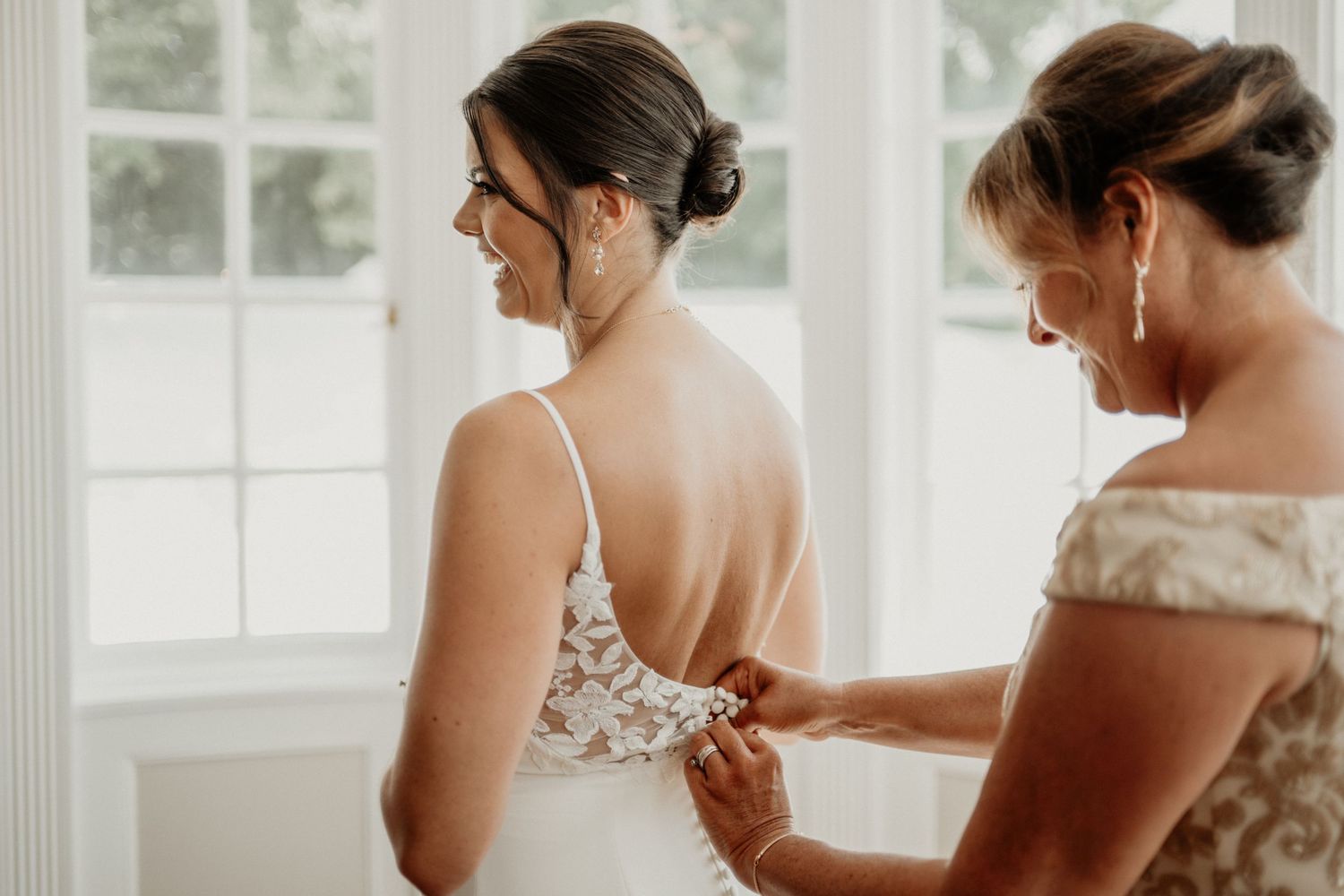 Bride in lace wedding gown getting dress fastened by bridesmaid in bright white room before ceremony.