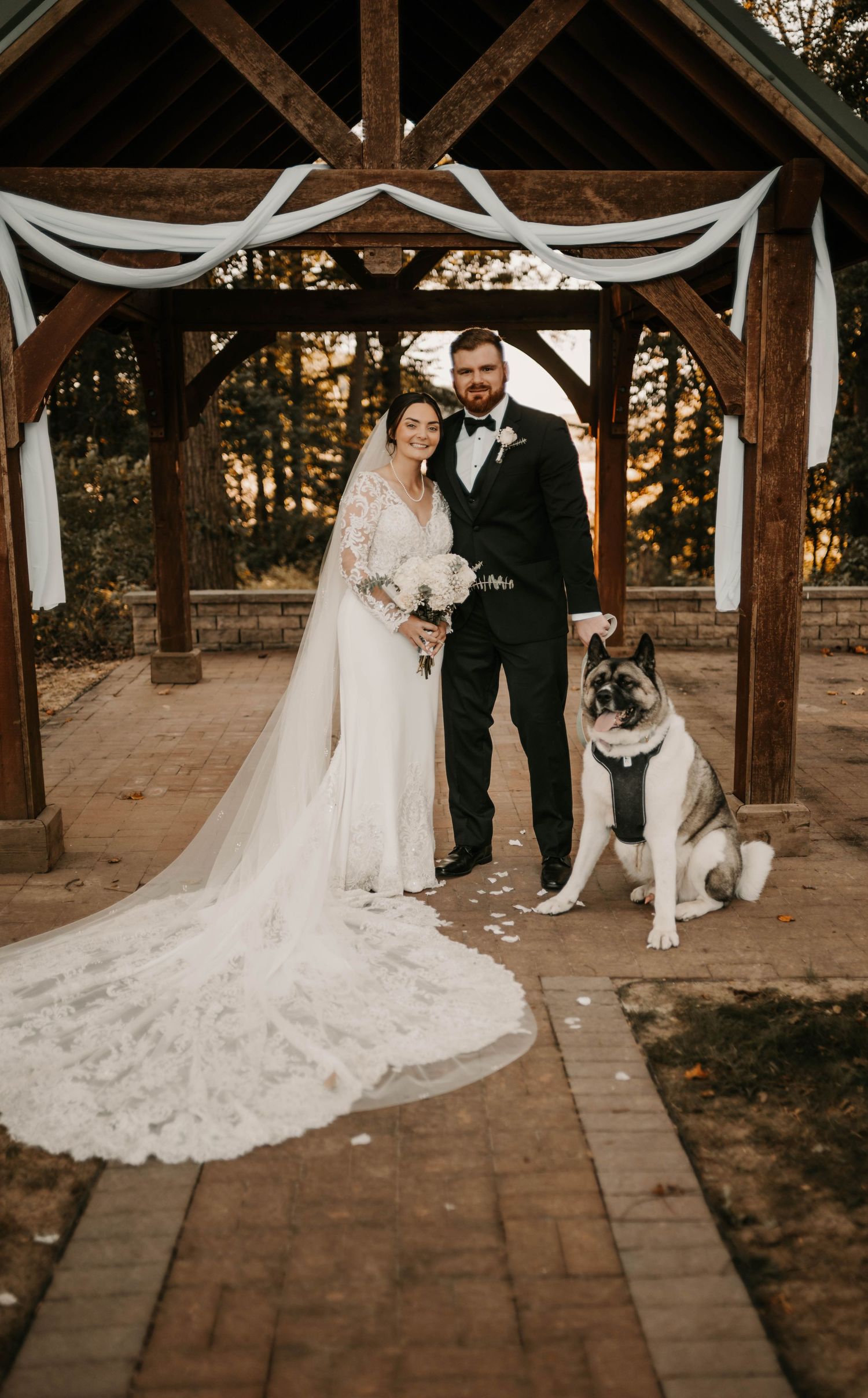 Bride and groom pose with their dog under a wooden gazebo at golden hour wedding ceremony.