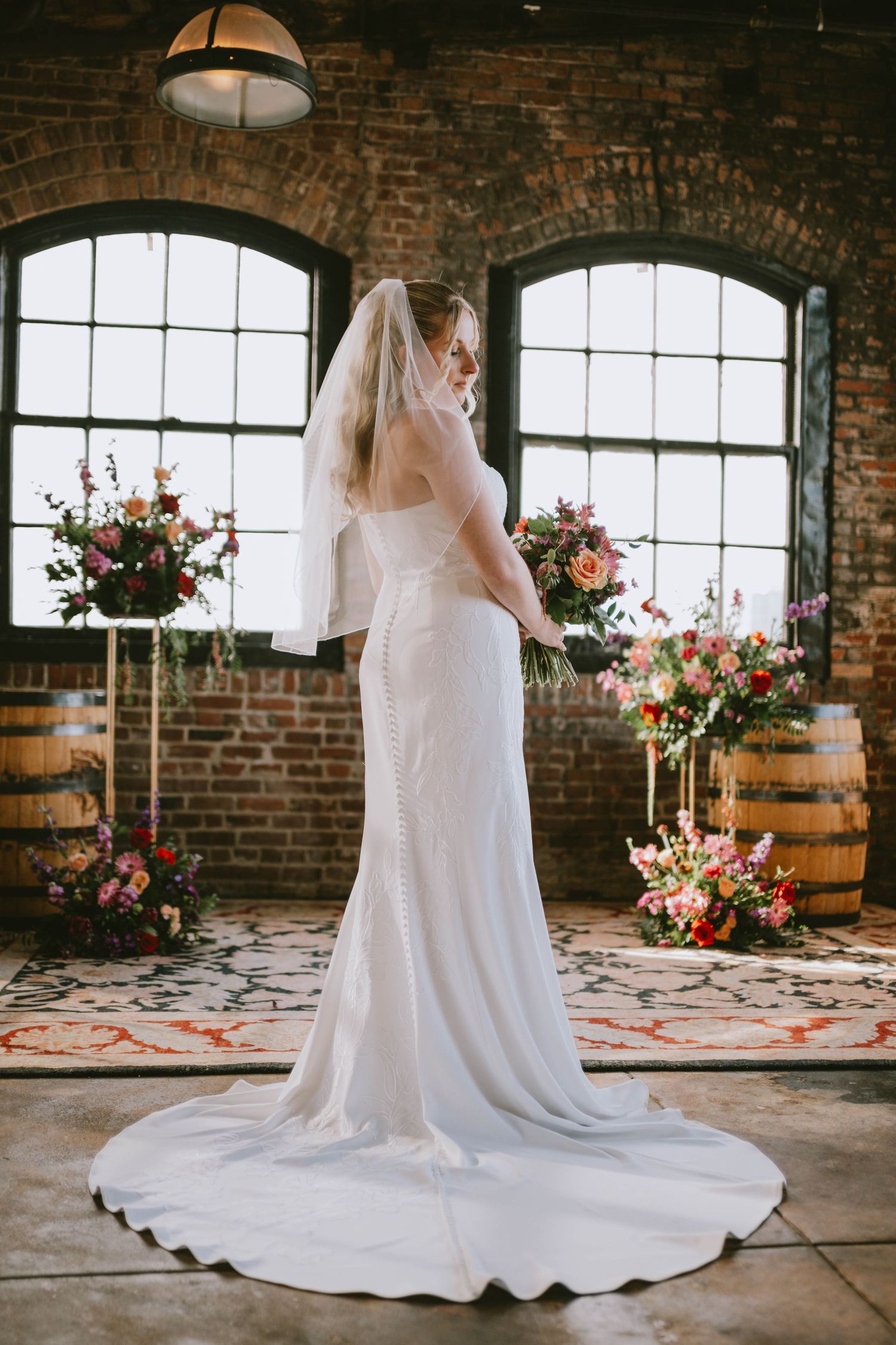 Bride in a fitted white lace gown with veil stands in rustic brick venue surrounded by vibrant floral arrangements.