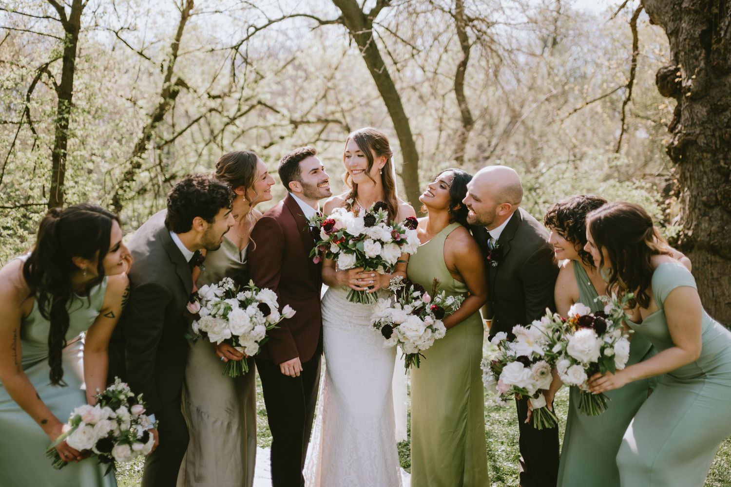 Wedding party celebrating outdoors in spring, bride and groom surrounded by bridesmaids in sage green and groomsmen in gray.