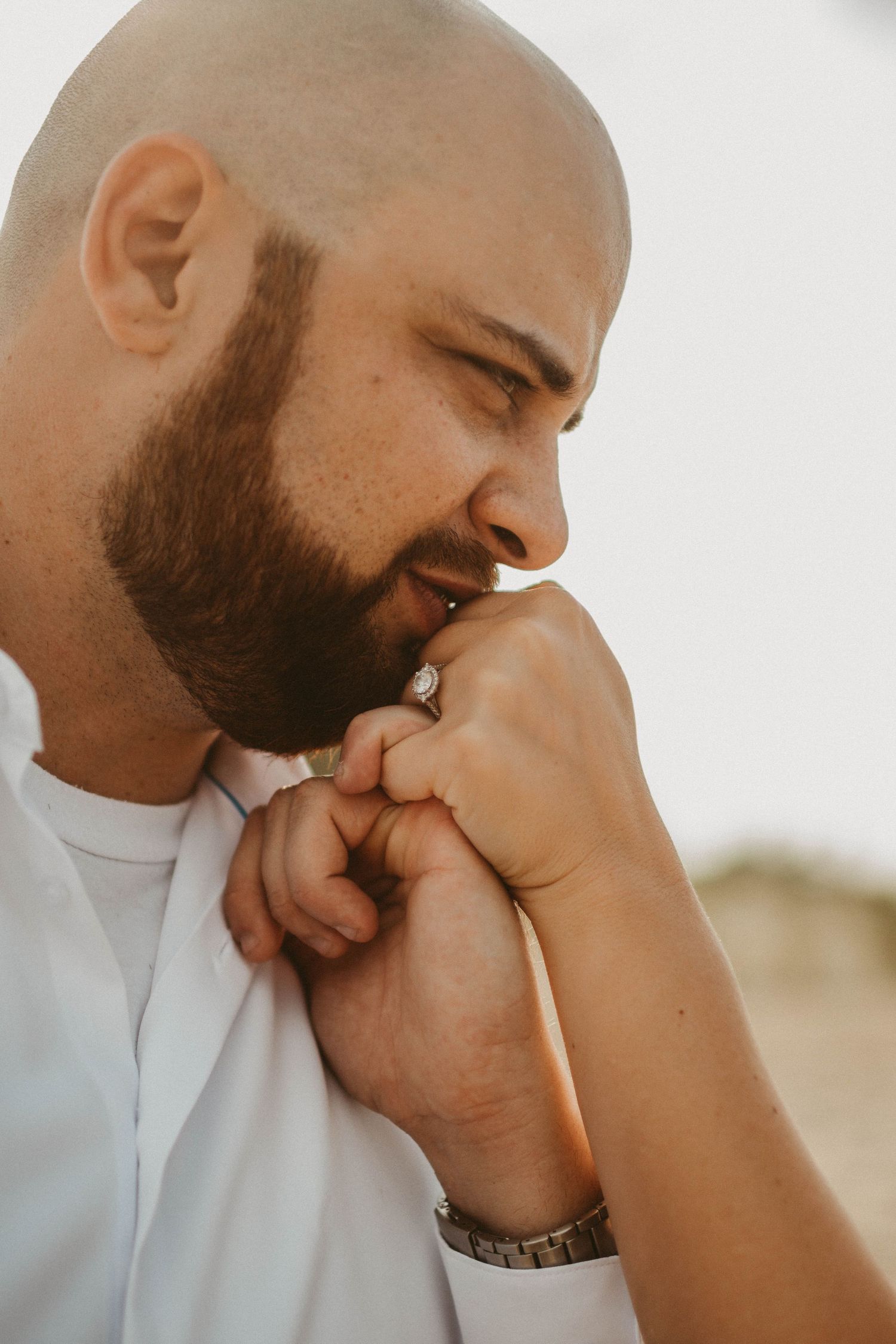 Bald bearded man in white shirt kissing a woman's hand wearing a ring and silver watch outdoors.