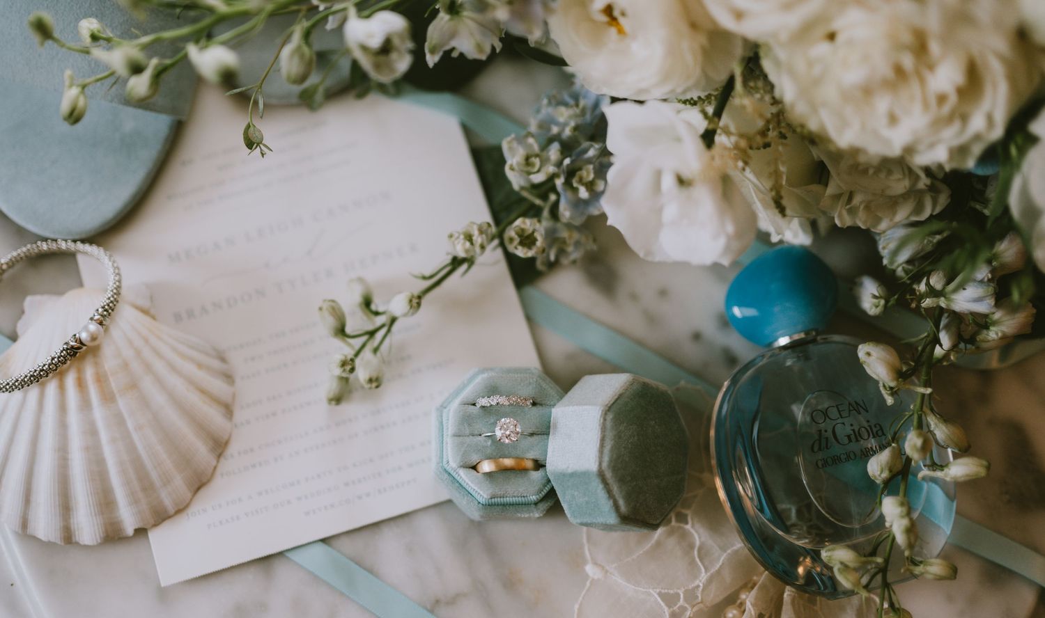 Elegant wedding flat lay with diamond ring in blue velvet box, white floral bouquet, seashell, and perfume bottle.