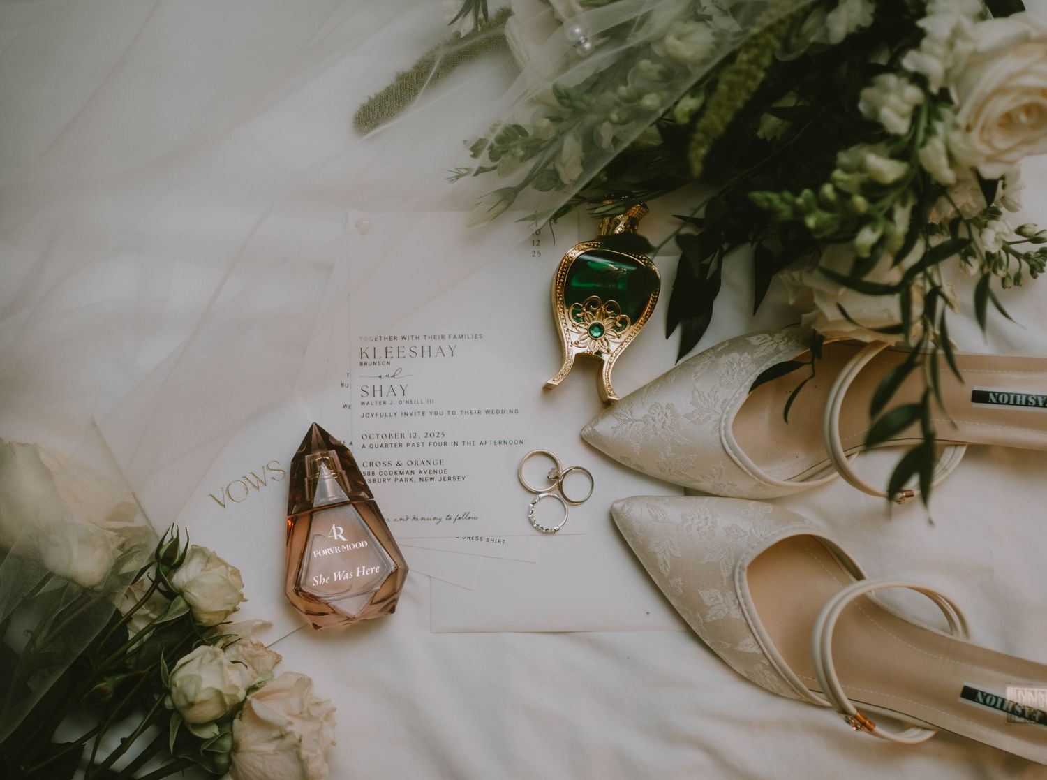 Elegant wedding flat lay with gold heels, rings, perfume bottles, green gemstone, and floral bouquet on white fabric.