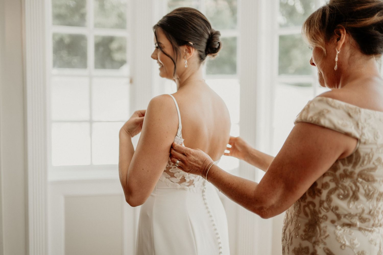 Mother helping bride button up elegant backless wedding gown near bright window on wedding day.