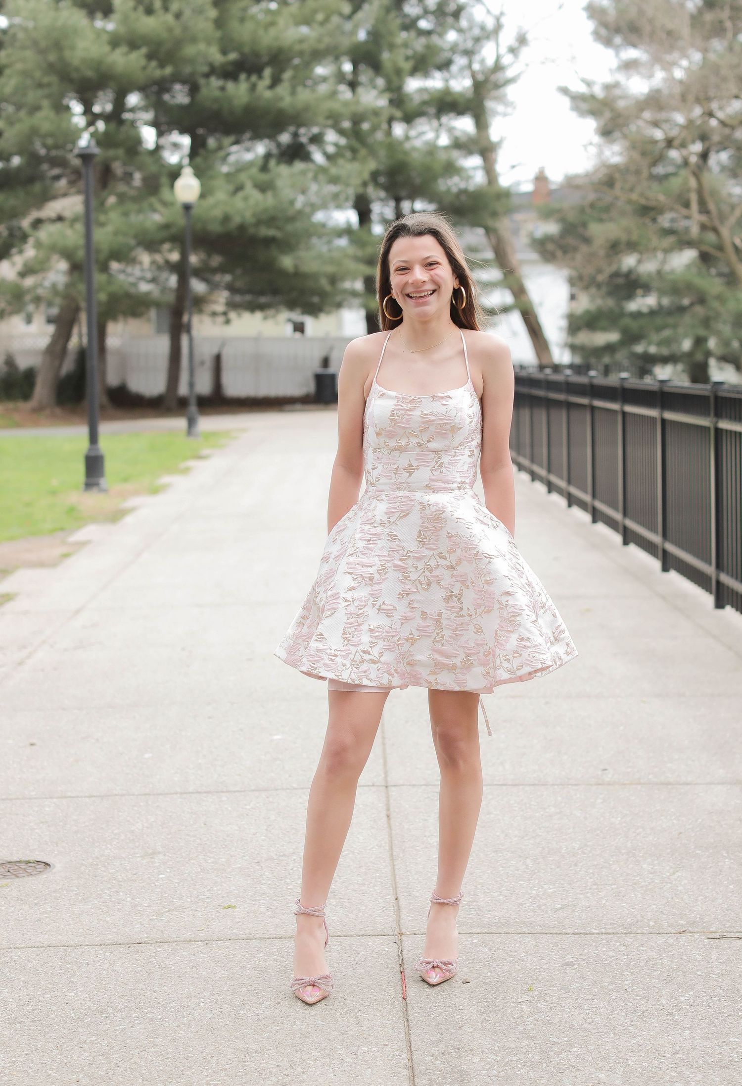 A sun-lit photo of someone in a white lace dress and pink heels standing on an outdoor walkway.
