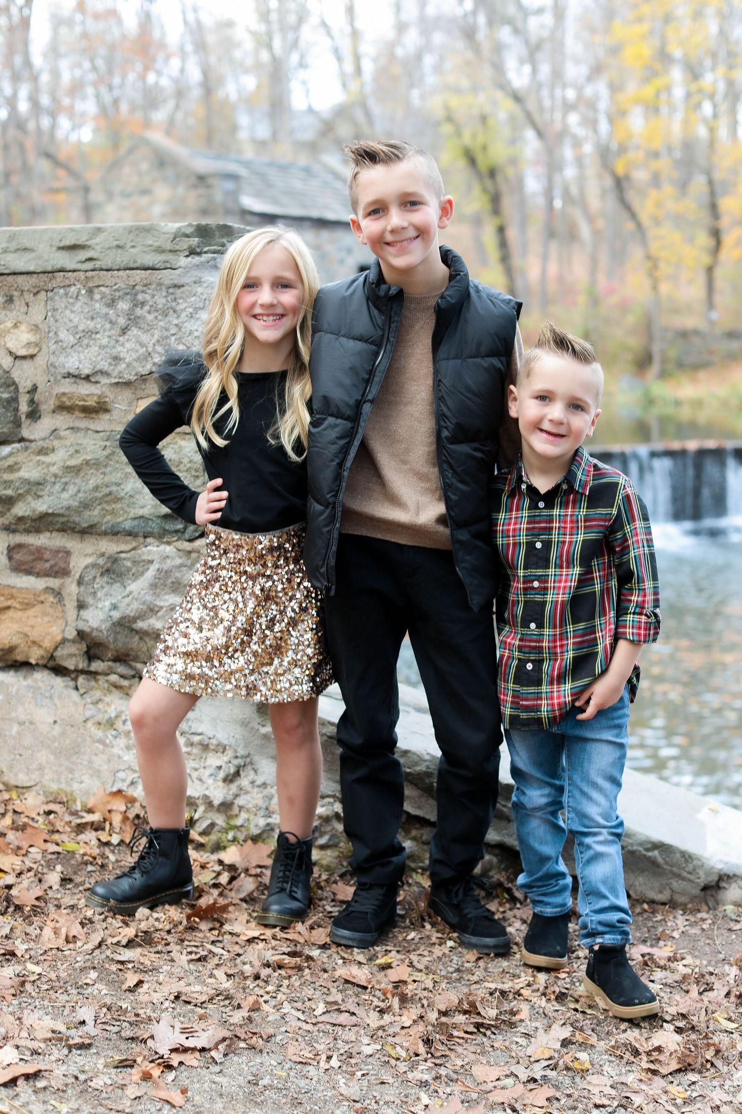 Three siblings pose together by stone wall near waterfall during autumn season.