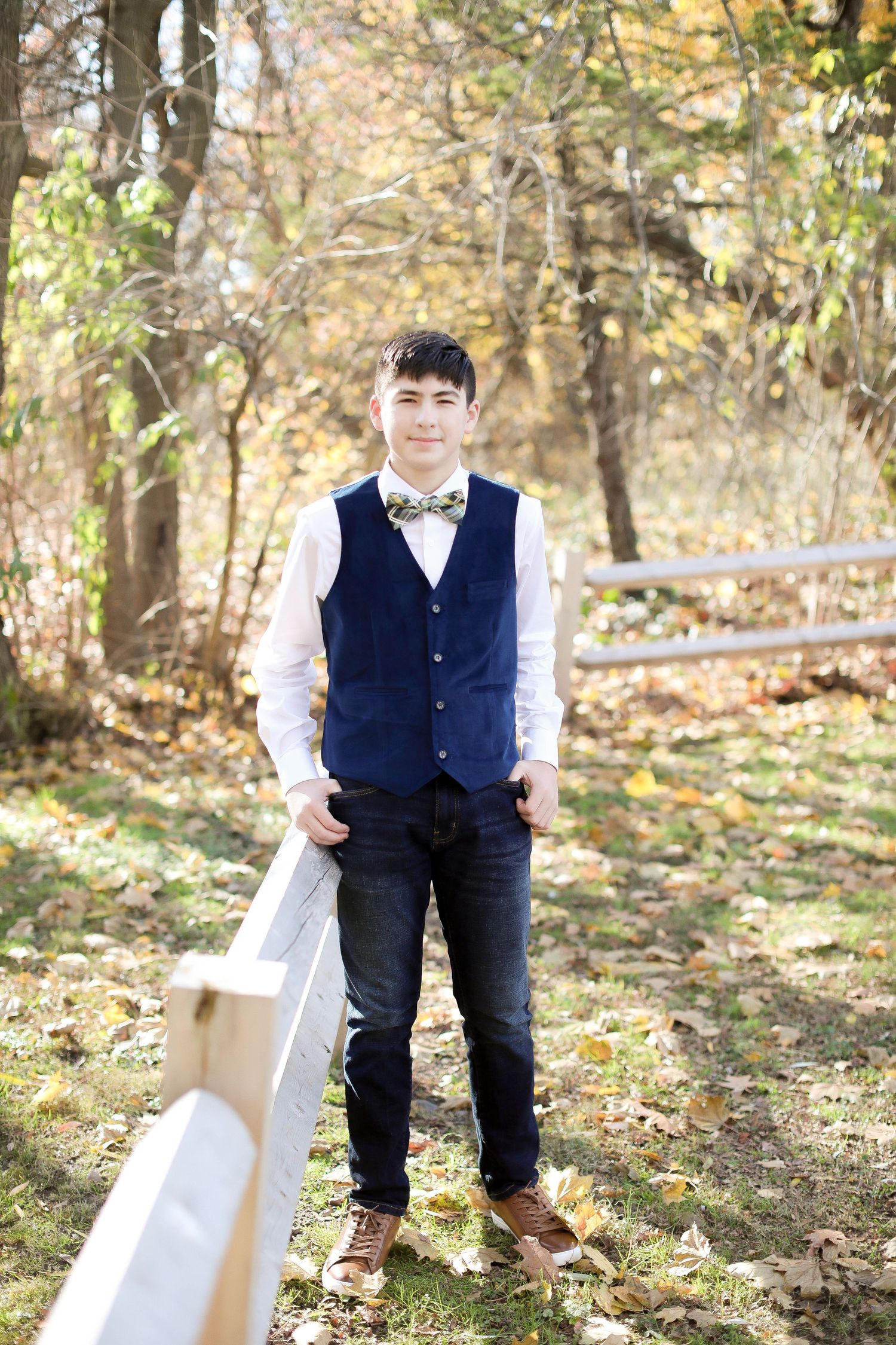 Student in navy vest and bowtie poses by white fence in autumn woodland setting.