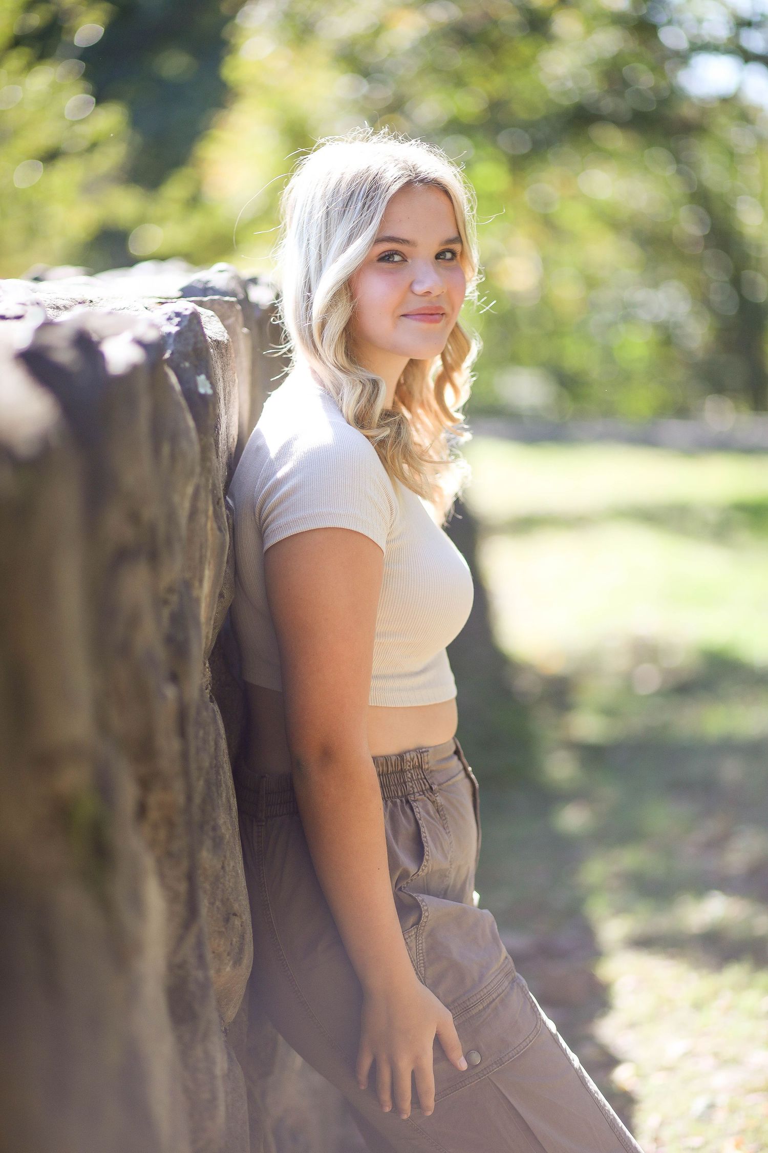 Woman in casual attire poses against stone wall in sunny outdoor setting.