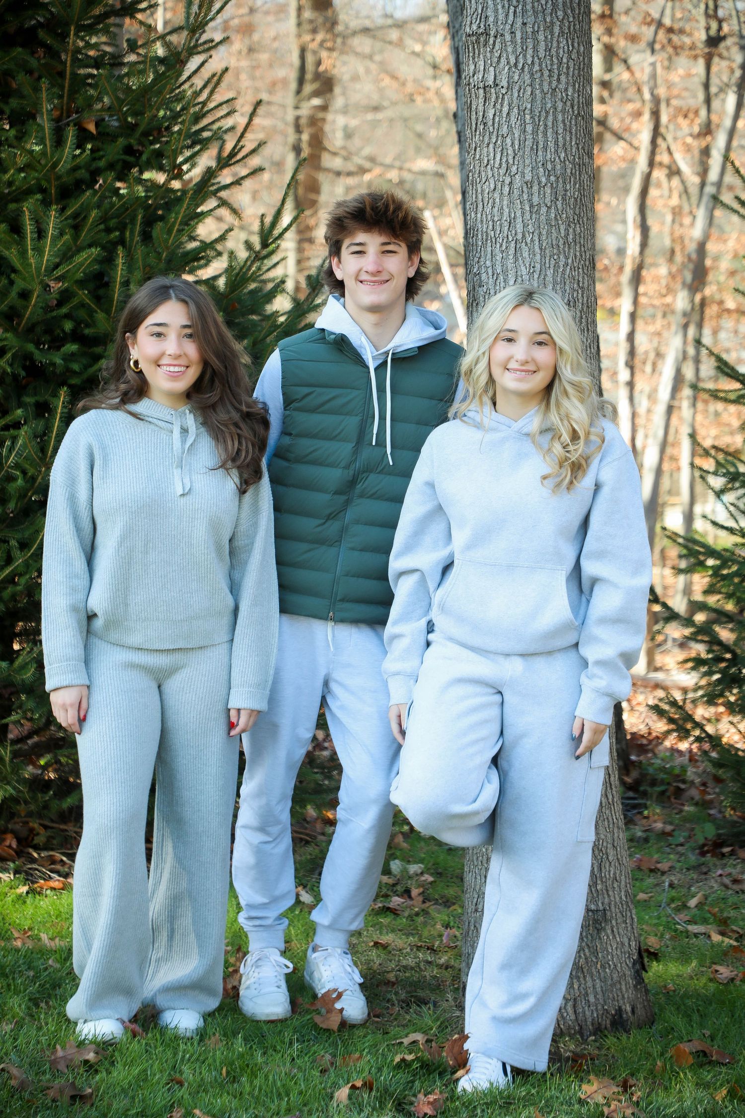 Three people in matching light gray athleisure wear posing by evergreen trees.