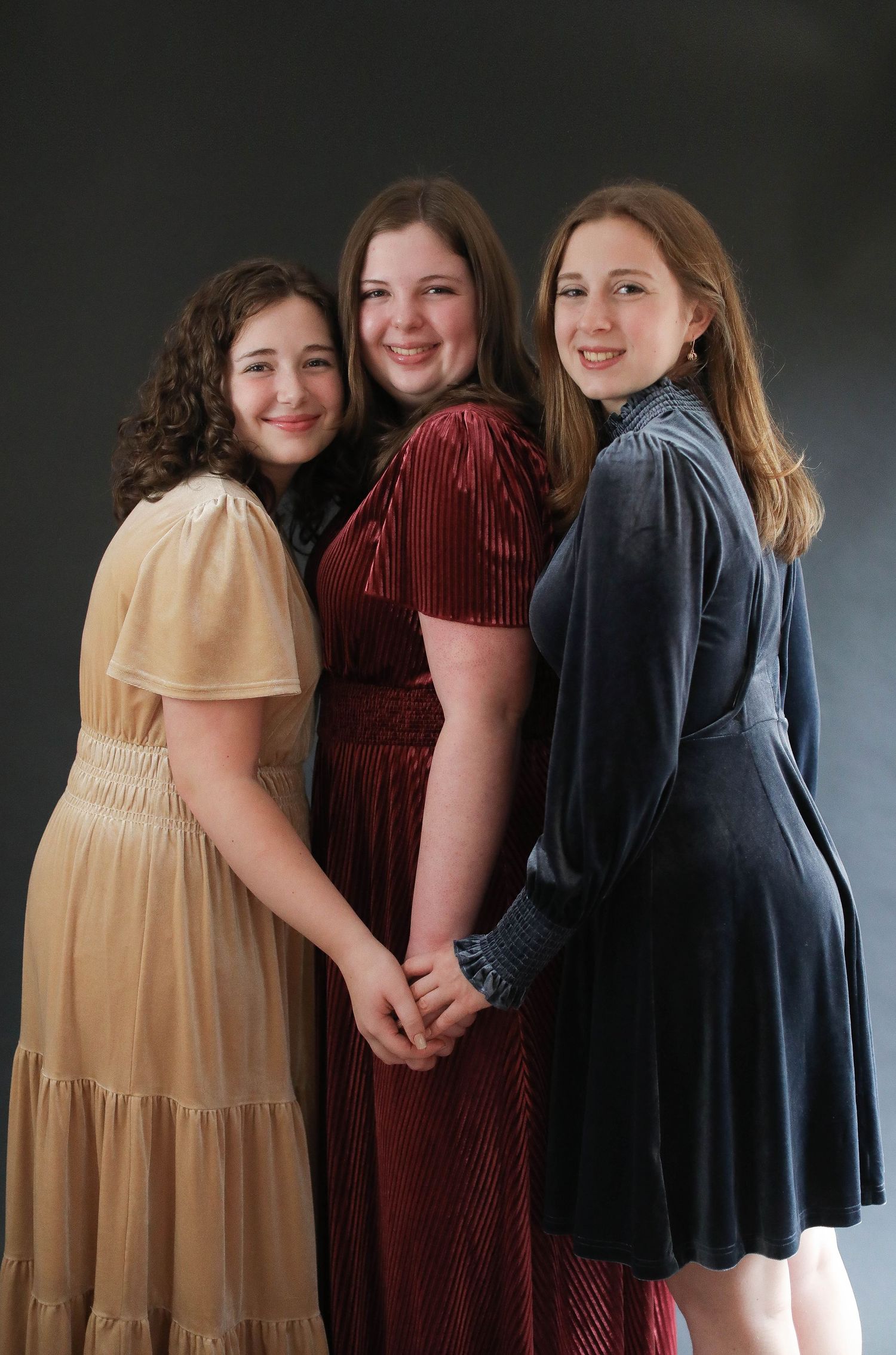 Three smiling friends pose together in dressy attire including a yellow, burgundy and navy velvet dress against dark backdrop.