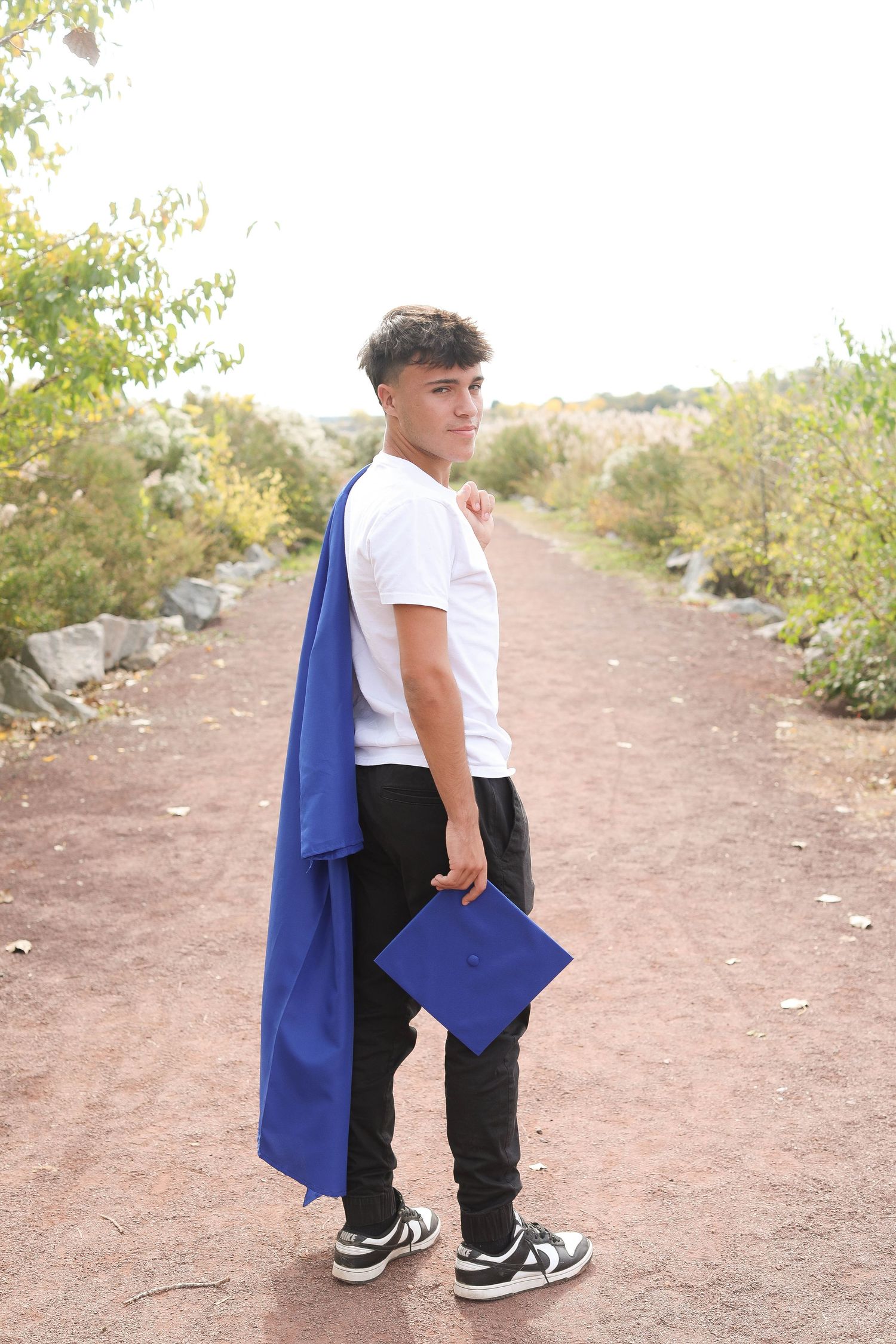 Graduate holding blue cap and gown while standing on a dirt path during sunset.
