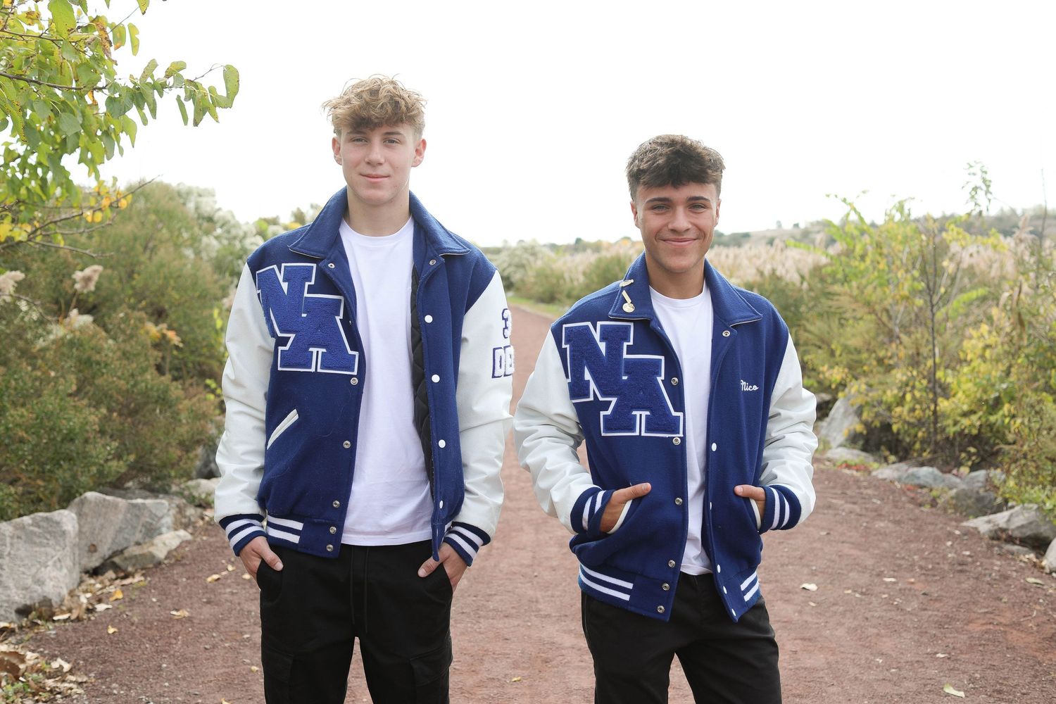 Two people wearing matching blue and white varsity letterman jackets standing on a dirt path.