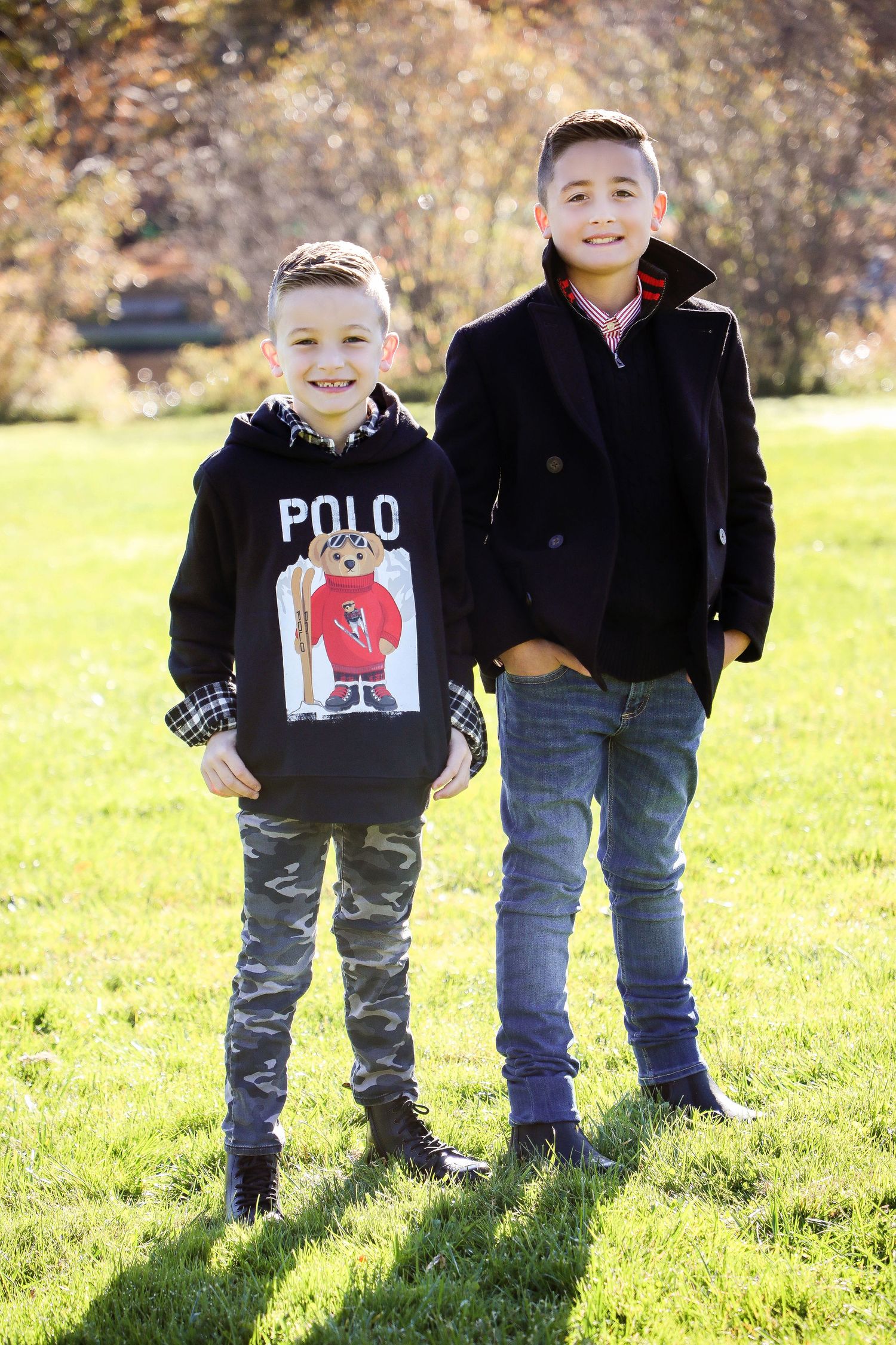 Two young boys in casual outerwear stand together on grass during fall season.