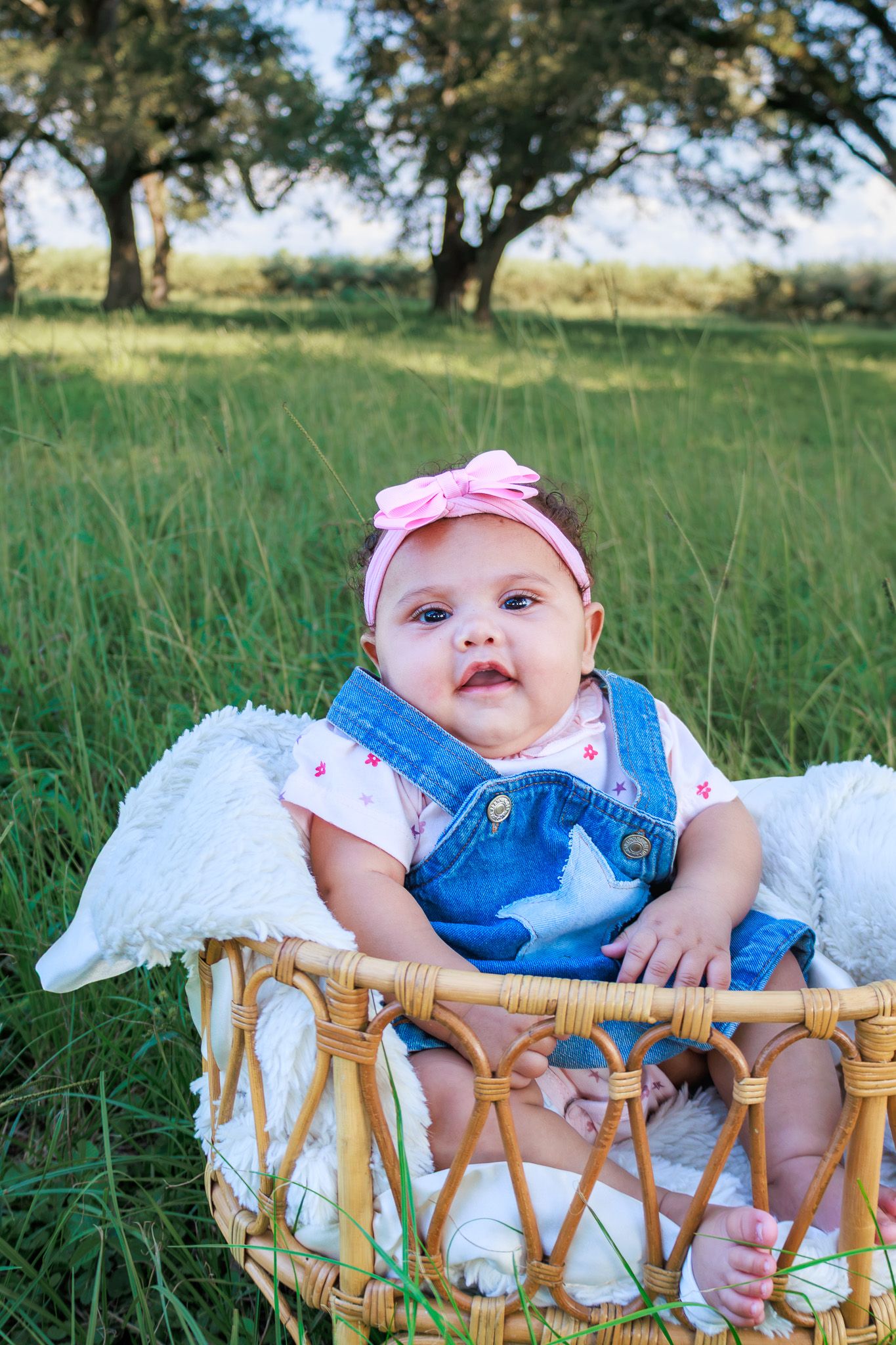 Adorable baby girl in denim overalls and pink bow headband sitting in a wicker basket outdoors in green field.