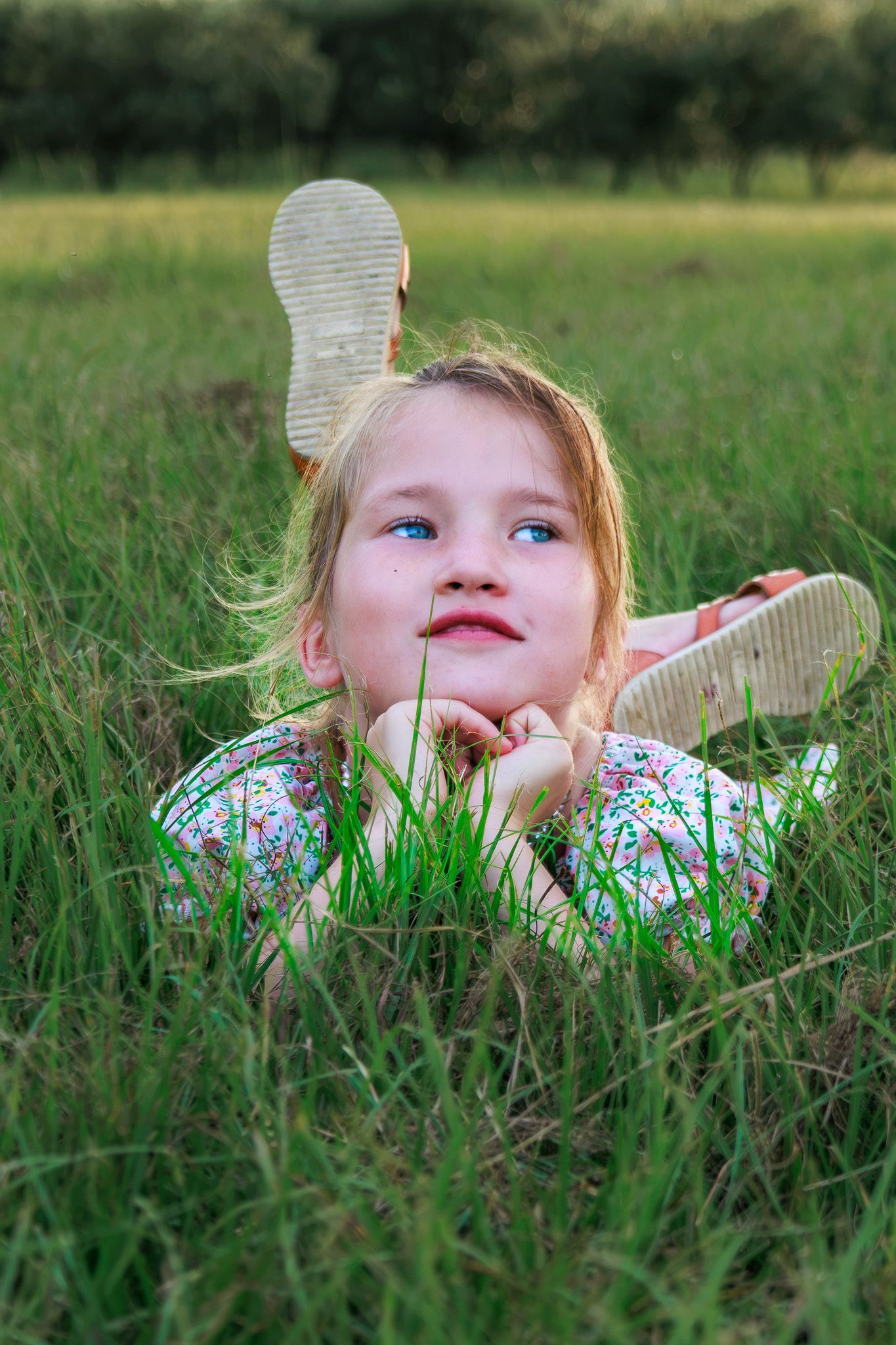 A blonde girl in a floral dress lies in green grass, resting her chin on her hands, looking up dreamily.