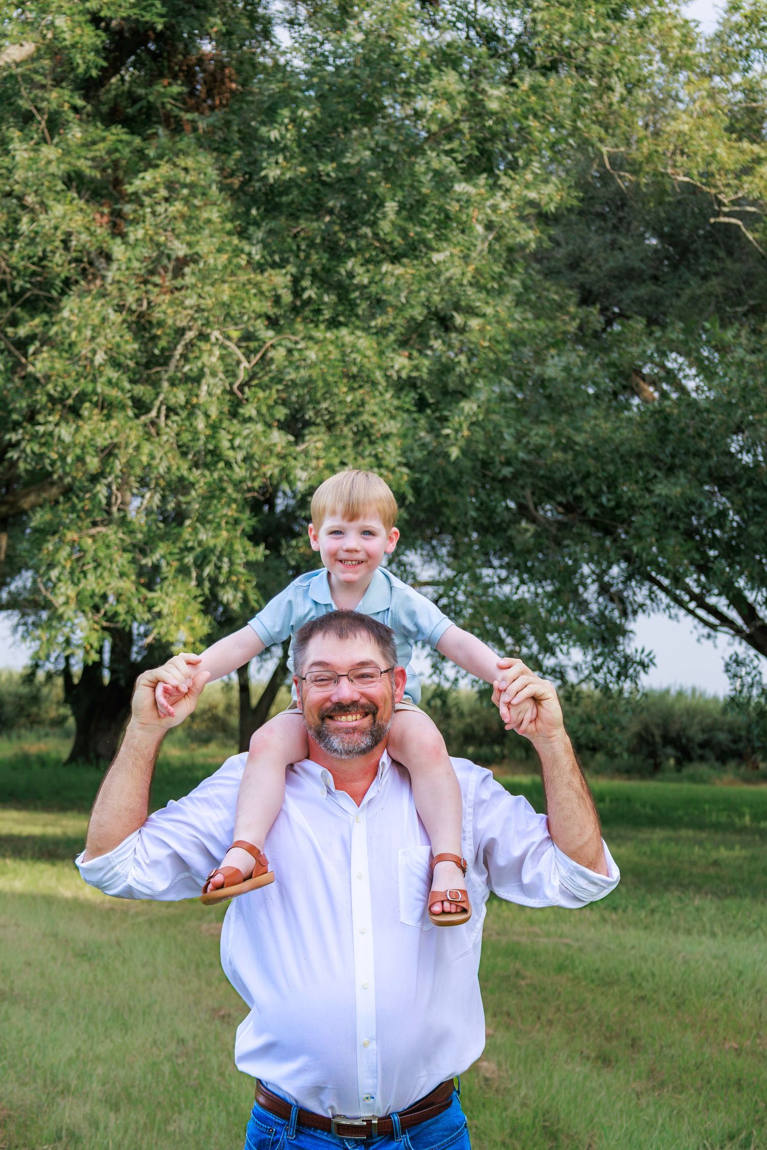 A smiling man in a white shirt carries a happy toddler boy on his shoulders outdoors in a green park.