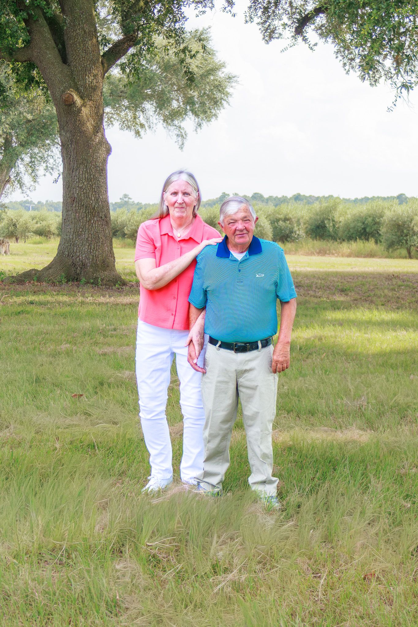 Elderly couple smiling together in a green field under a large oak tree on a sunny day.