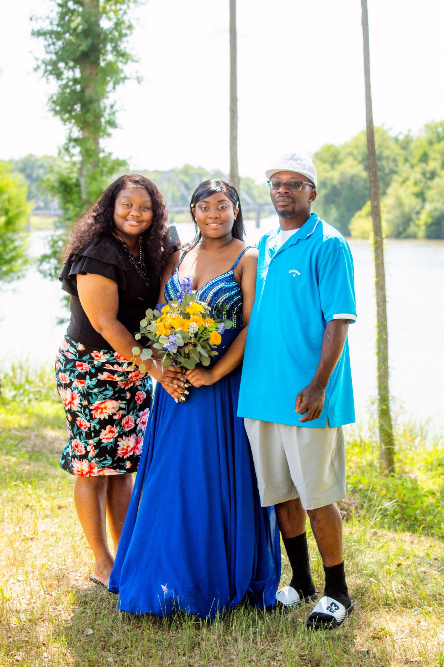 A prom girl in a blue gown poses with her parents outdoors near a river, holding a yellow bouquet.