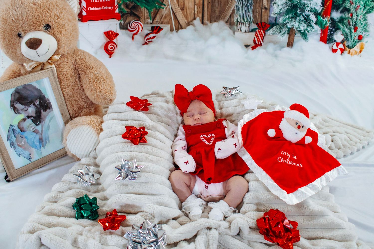 Newborn baby in red outfit lies on white blanket surrounded by Christmas decorations and gifts.