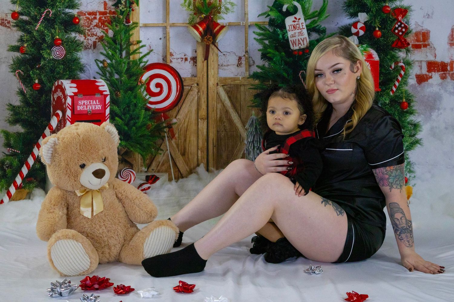 Woman in black pajamas sitting with a young child and large teddy bear in front of a Christmas holiday backdrop.