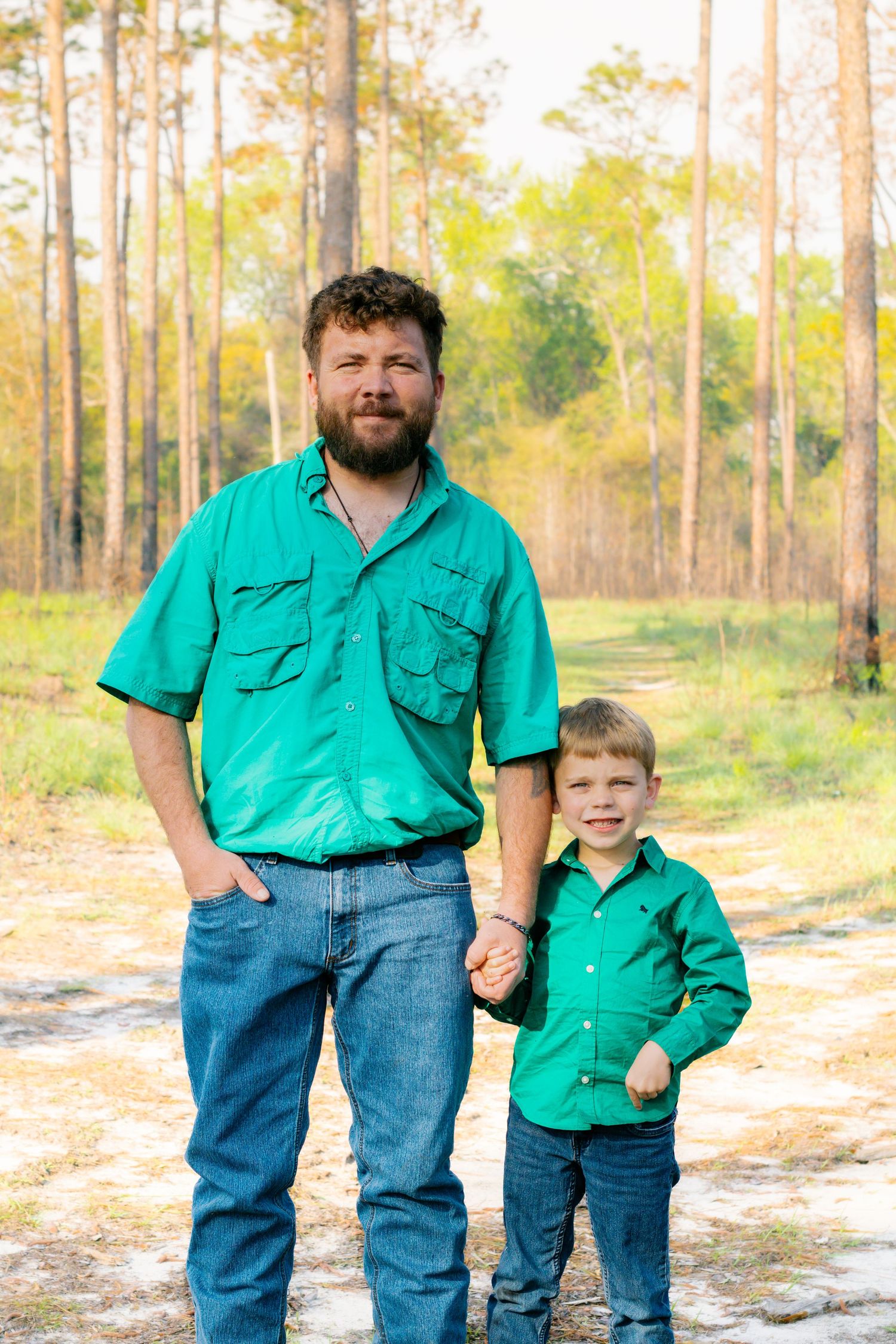 A father and son in matching green shirts hold hands while standing on a dirt path in a pine forest.