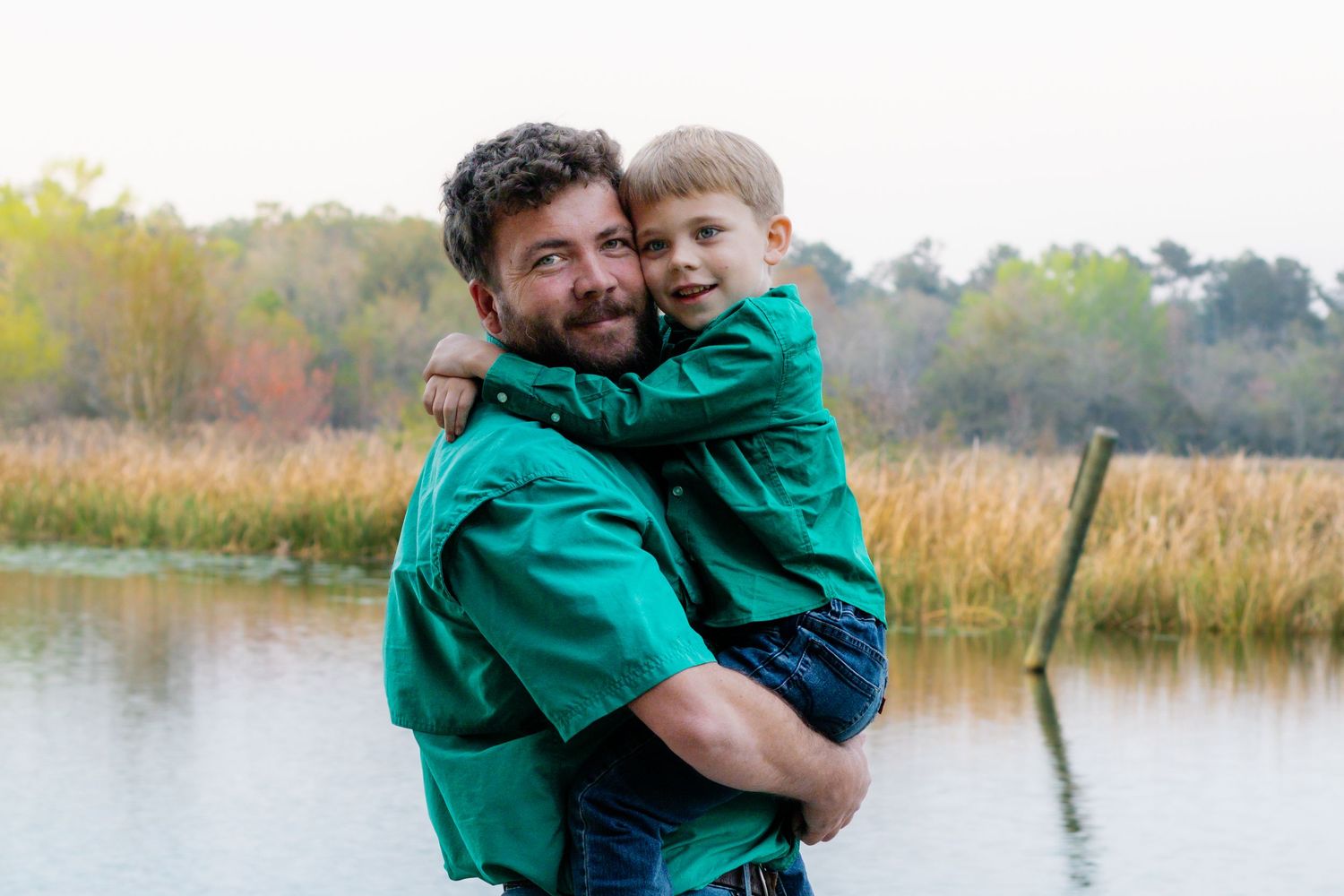 Bearded father in green shirt holds smiling young son near a scenic lake with tall grass.