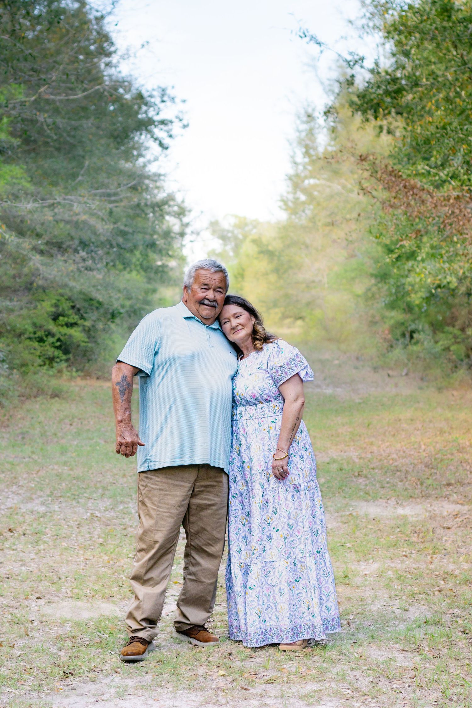 Older couple embracing on a dirt path surrounded by lush green trees, smiling at camera.