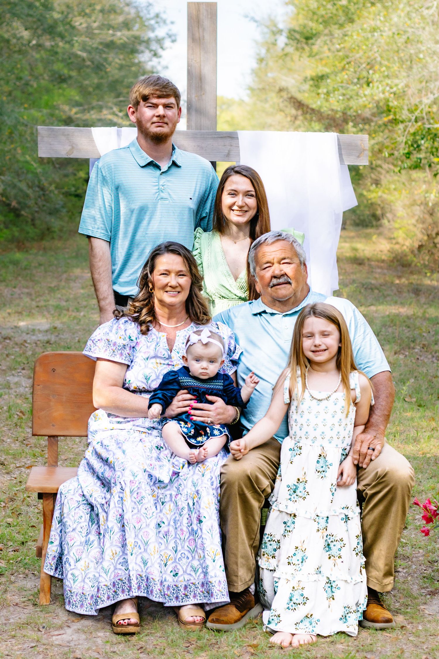 Multi-generational family of six posing in front of a wooden cross in a natural outdoor setting.