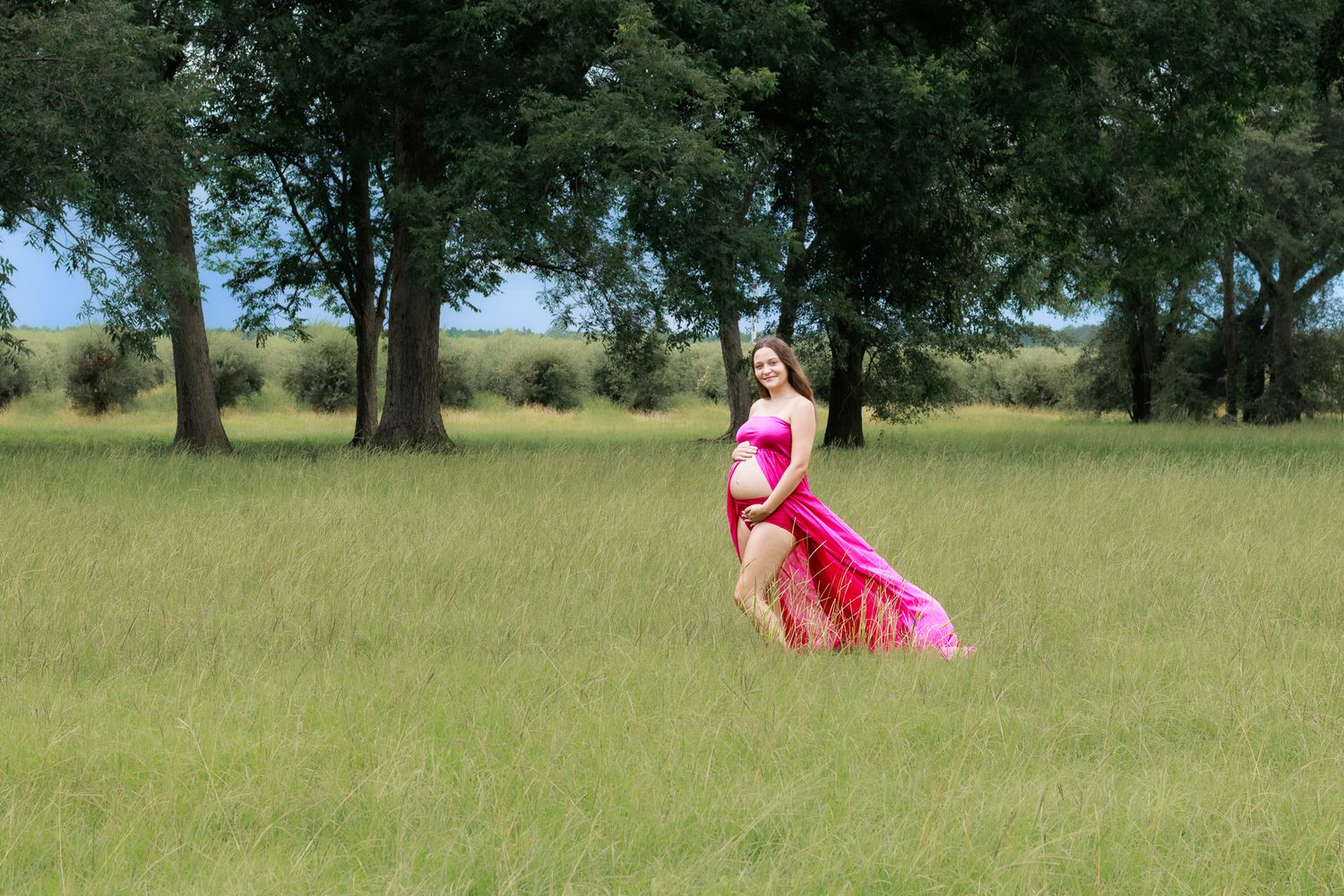 Pregnant woman in flowing pink dress posing in green field with trees.