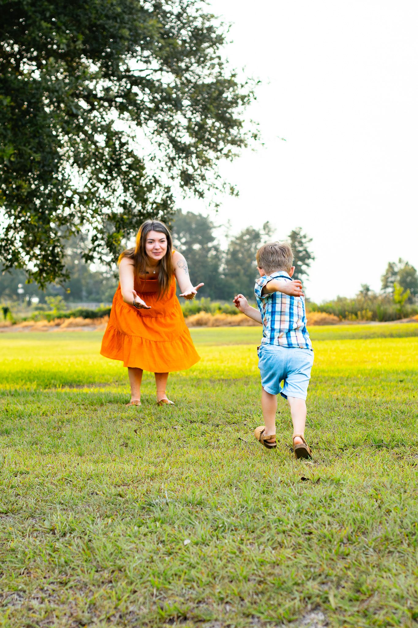 A smiling woman in an orange dress reaches out to a young boy running to her on a lush green lawn near trees.