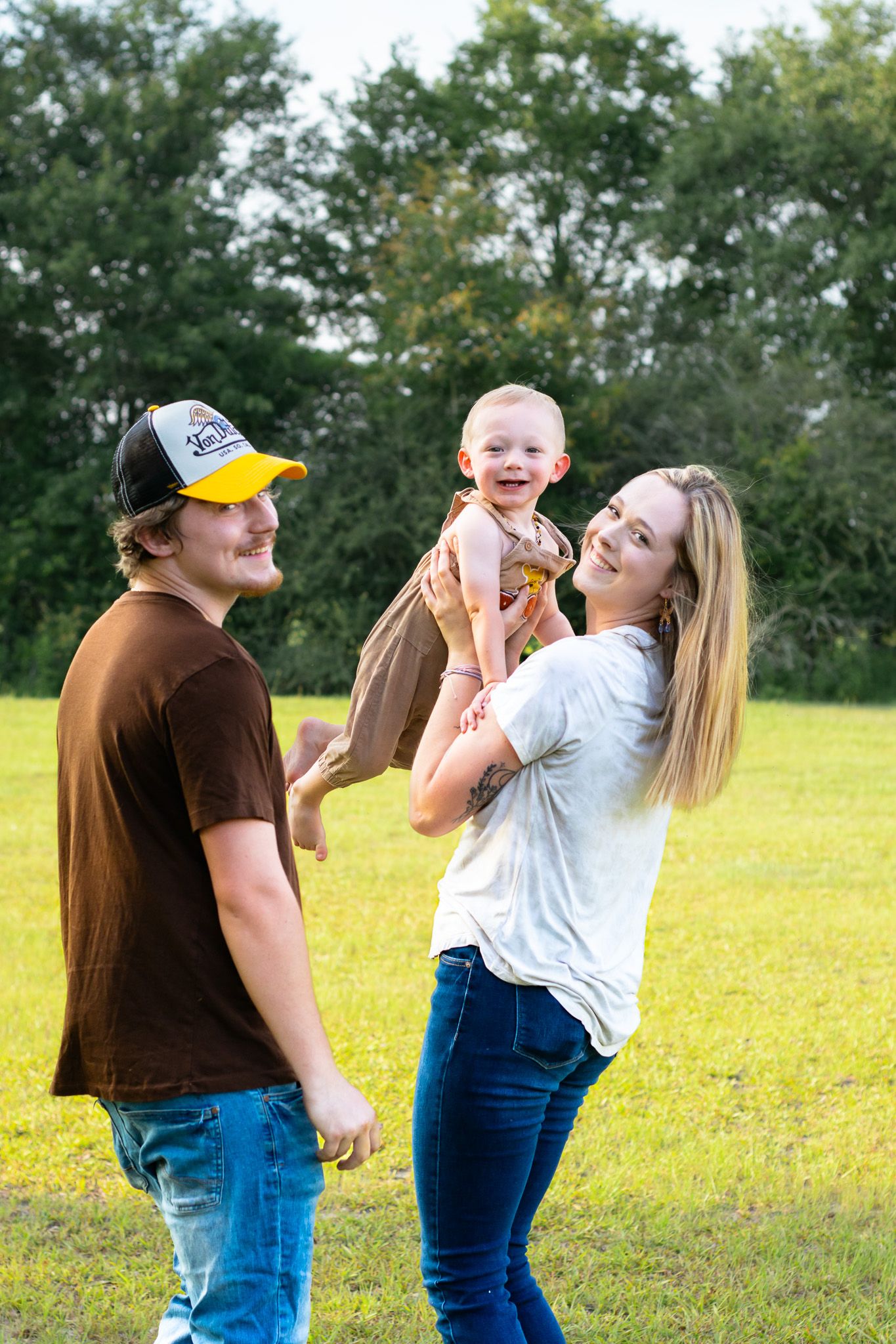 Young family posing outdoors on green grass, parents smiling as mother lifts happy baby in brown outfit.