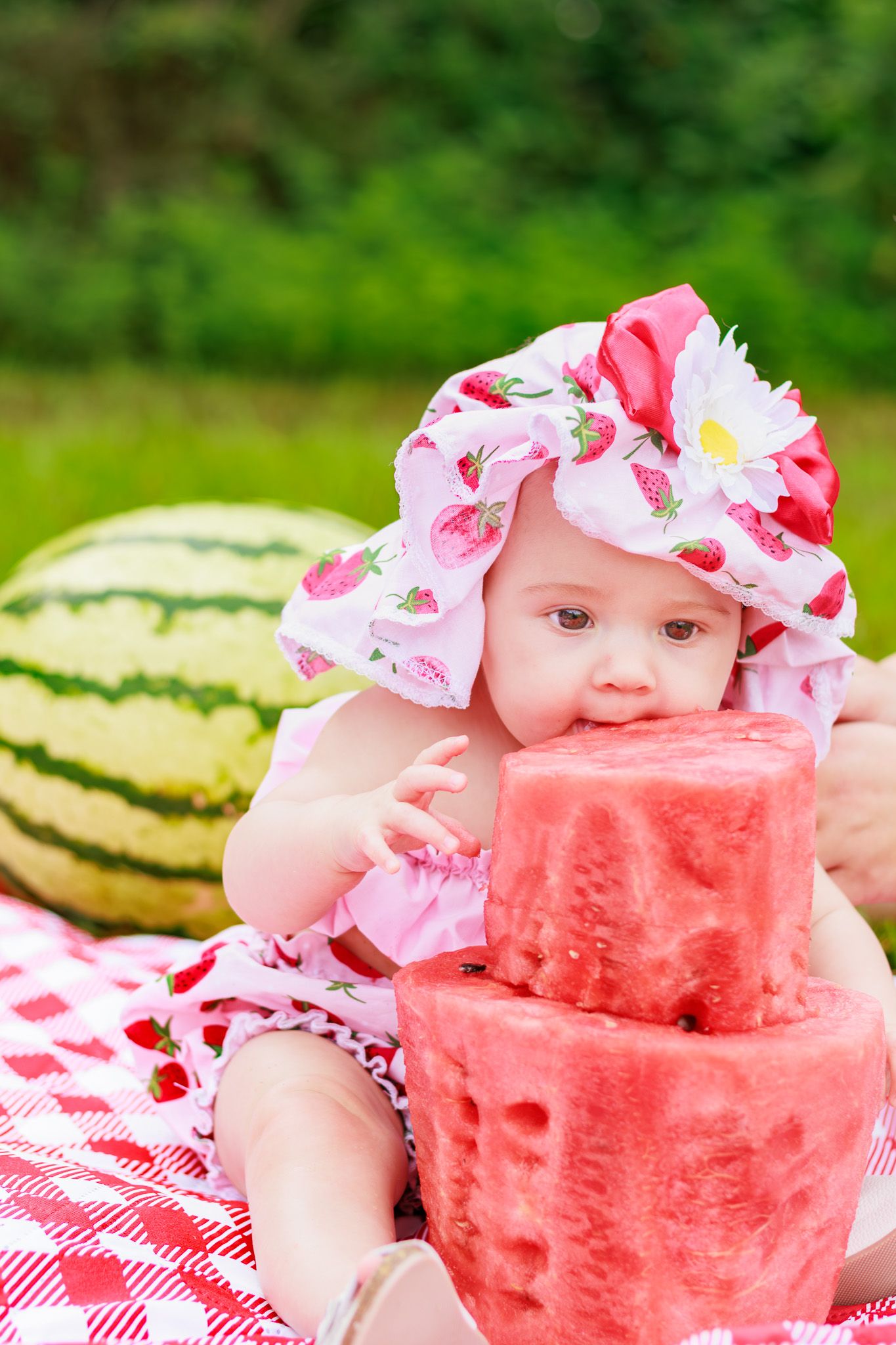 Adorable baby in a strawberry print hat biting into a large slice of watermelon on a red checkered blanket outdoors.